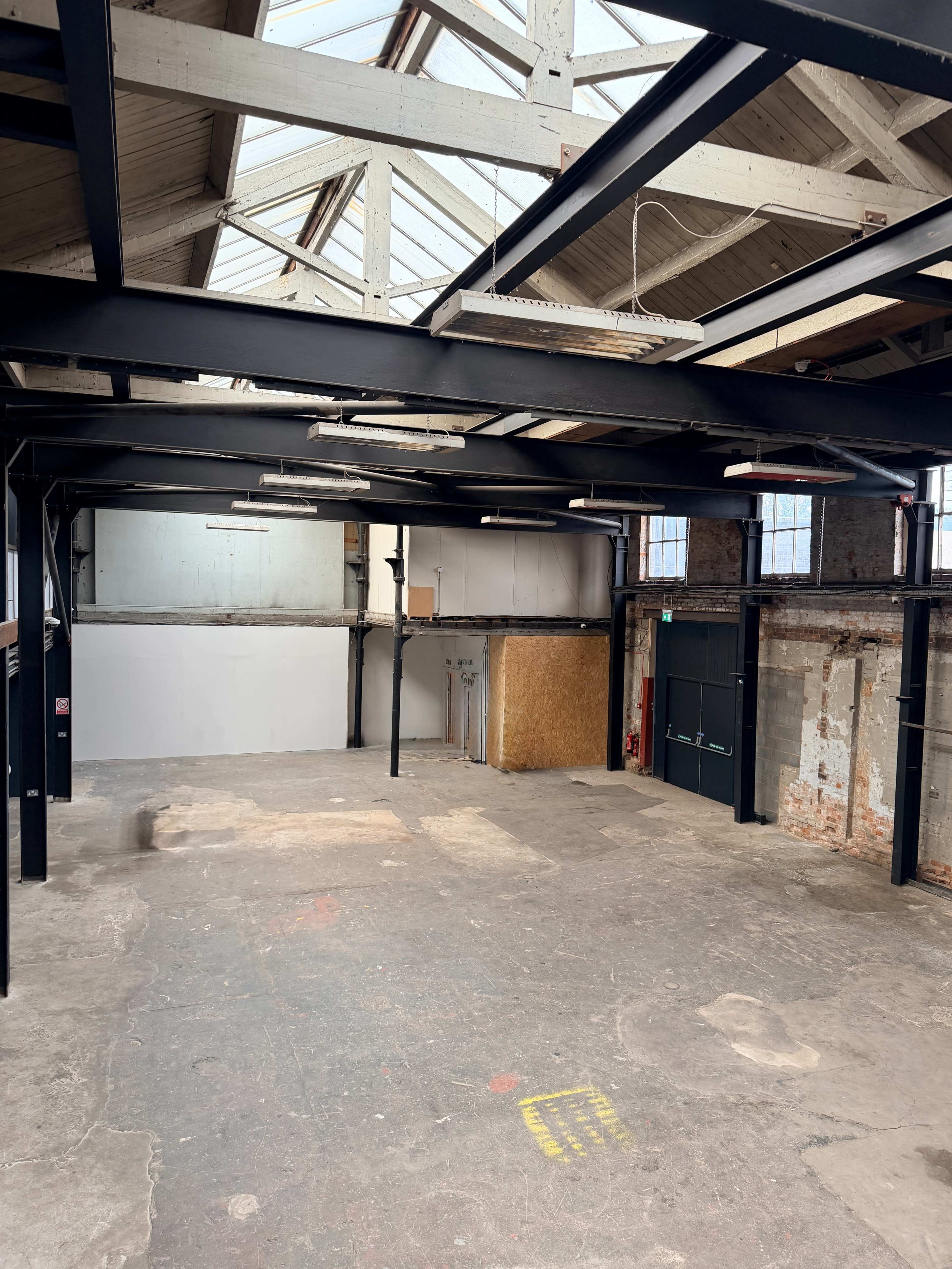 The image shows an industrial-style warehouse interior with exposed beams, a concrete floor, and large windows allowing natural light to enter.