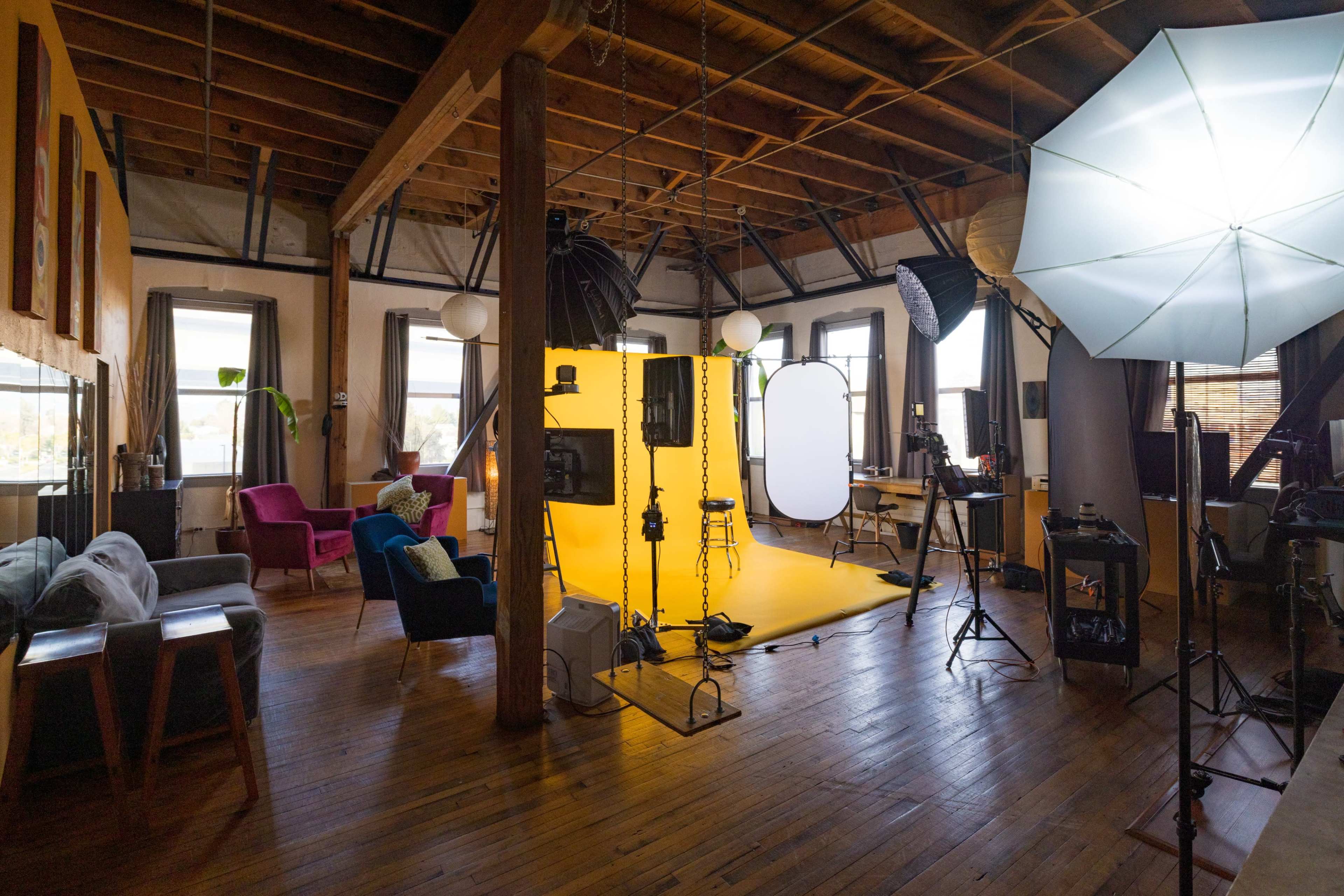 The image shows a spacious photography studio with a bright yellow backdrop, various lighting equipment, and seating areas.