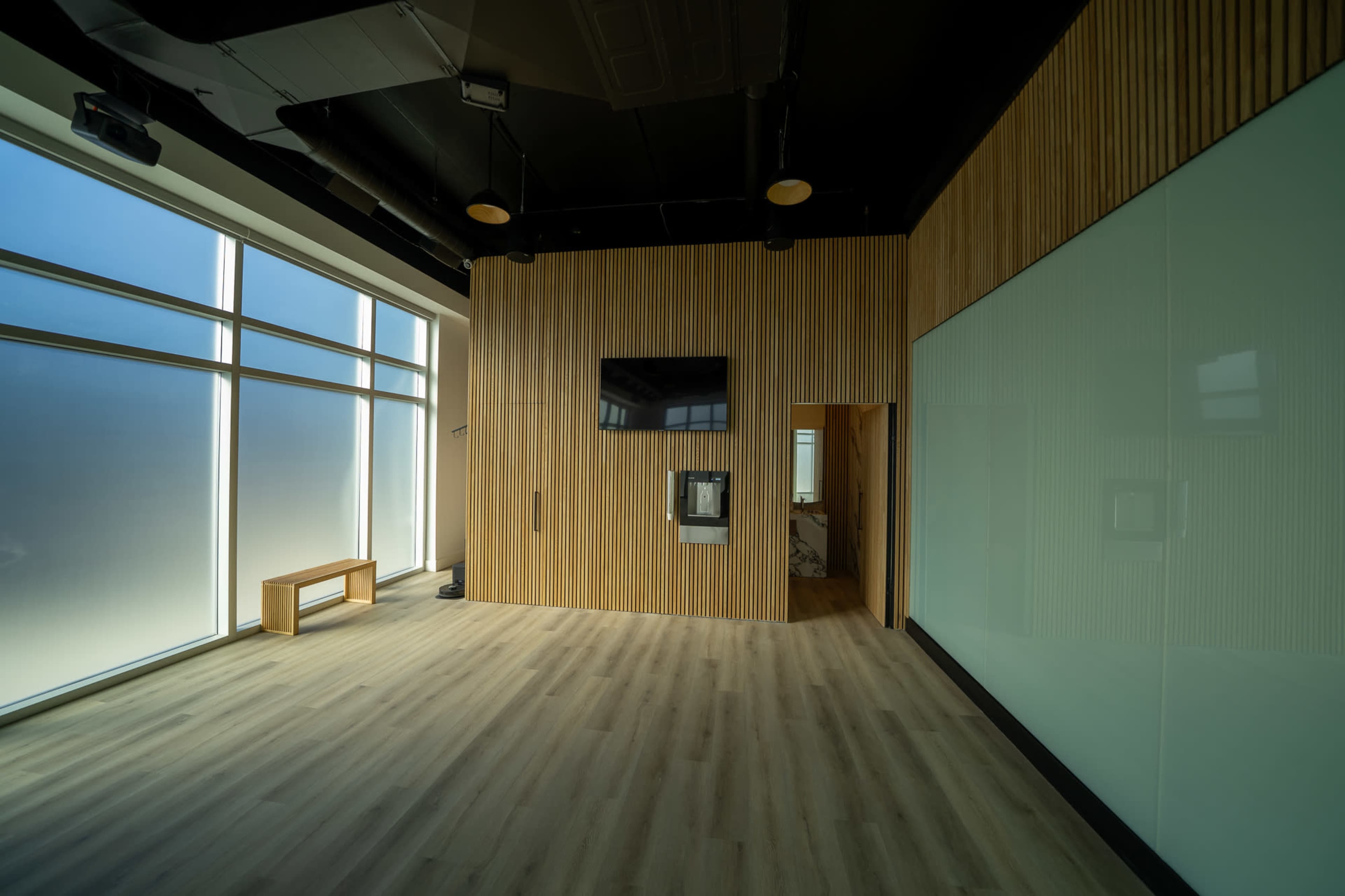 The image shows a modern, empty office space with wooden paneling, large windows, and a wall-mounted television.