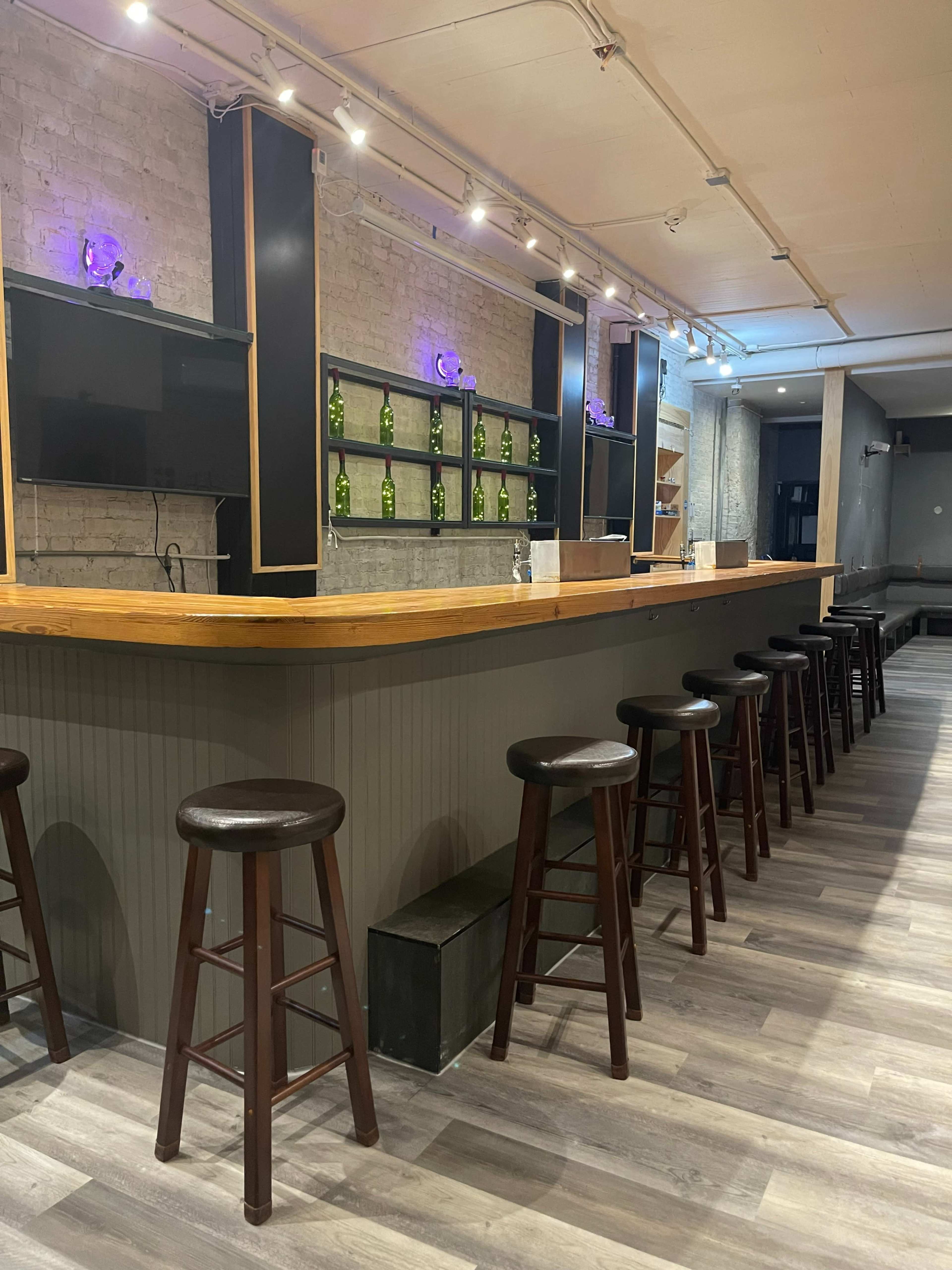 A modern bar area with a wooden countertop, several high stools, and a wall display of green bottles.