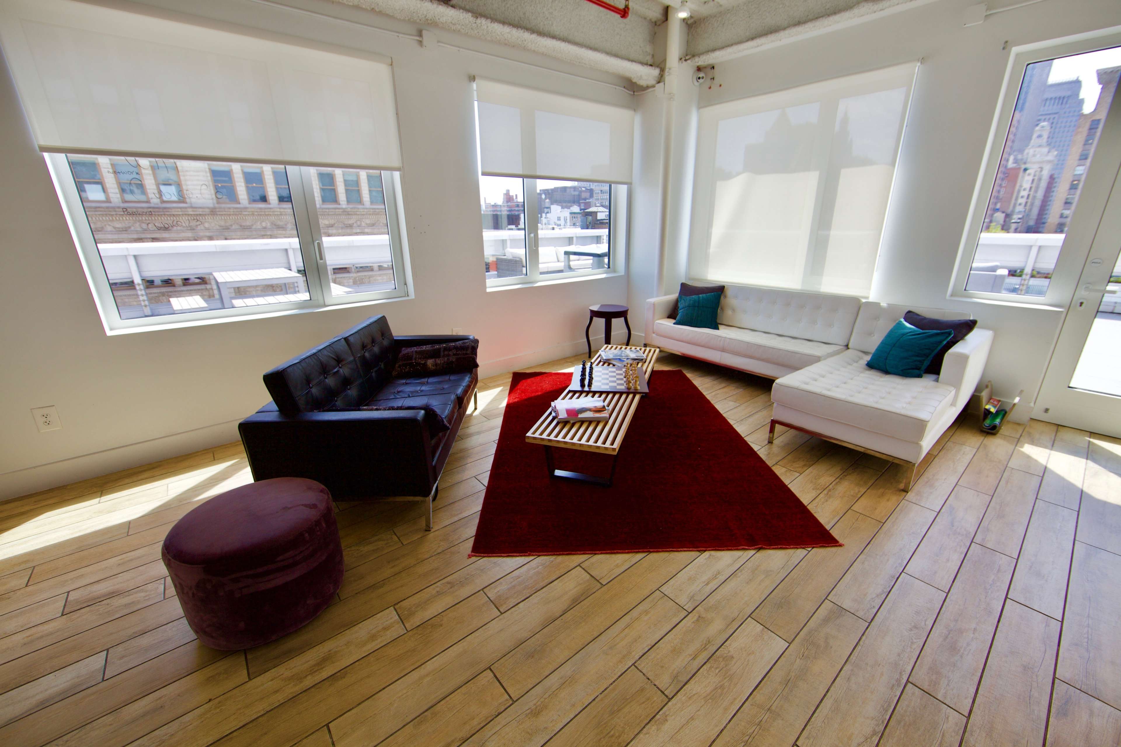 The image shows a bright, modern living room with large windows, a black leather sofa, a white sectional couch, a round ottoman, and a red area rug centered around a coffee table.