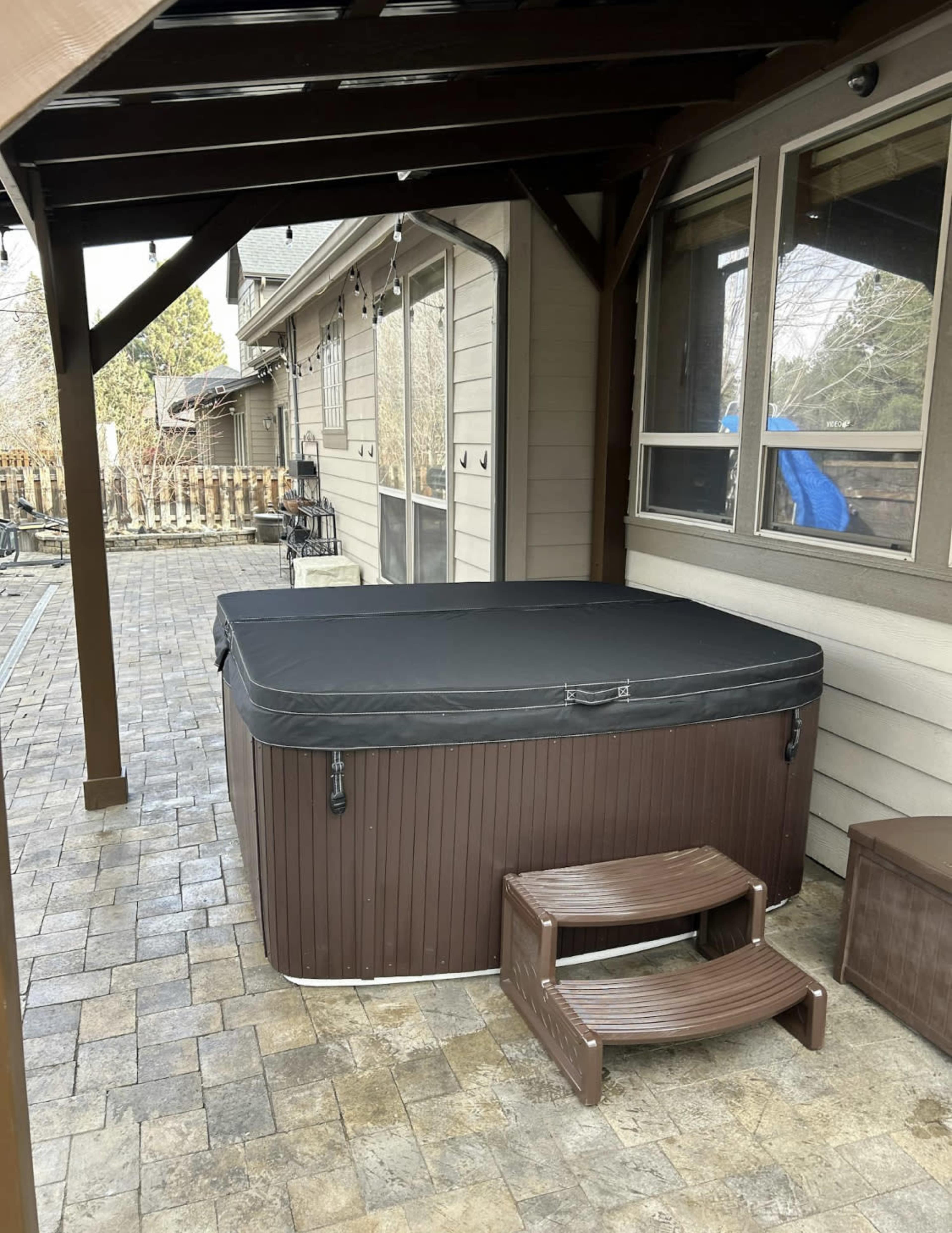 A hot tub with a black cover is placed on a stone patio under a wooden canopy next to a house.