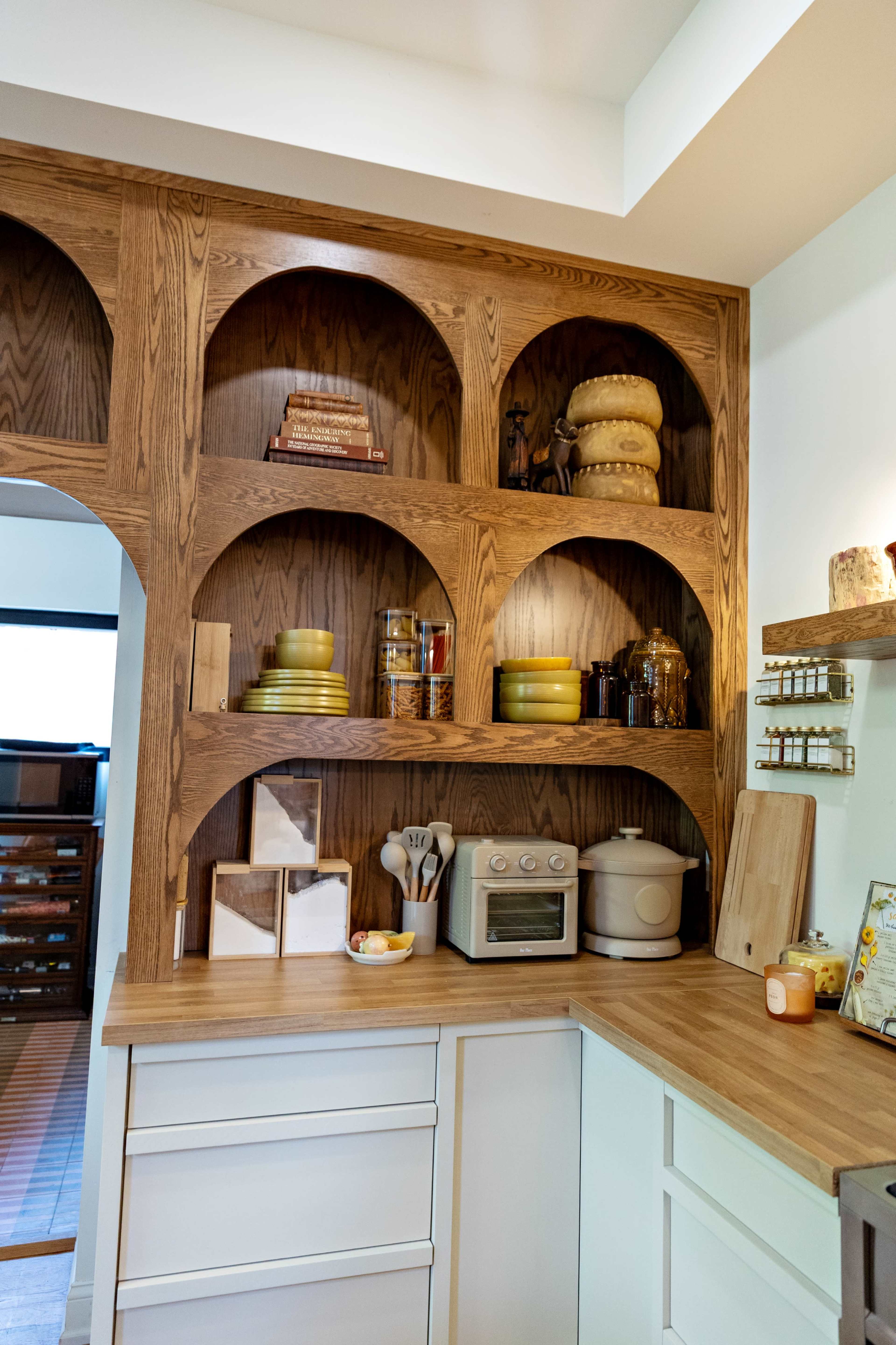 The image shows a kitchen shelf made of wooden arches, displaying various dishes, jars, and kitchen appliances on a countertop.
