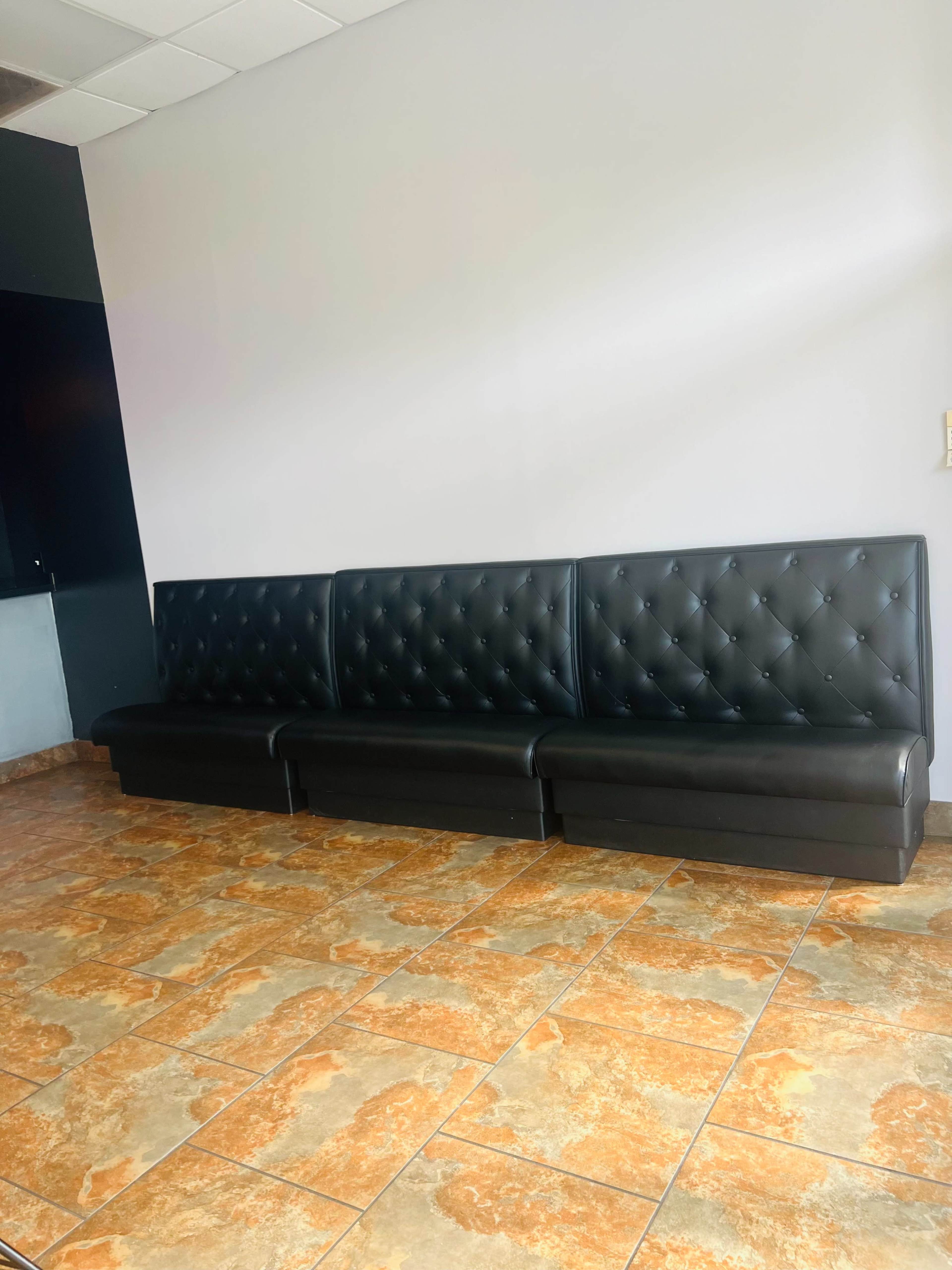 The image shows a long black leather upholstered bench against a light-colored wall, with a tiled floor in a pattern of orange and brown.
