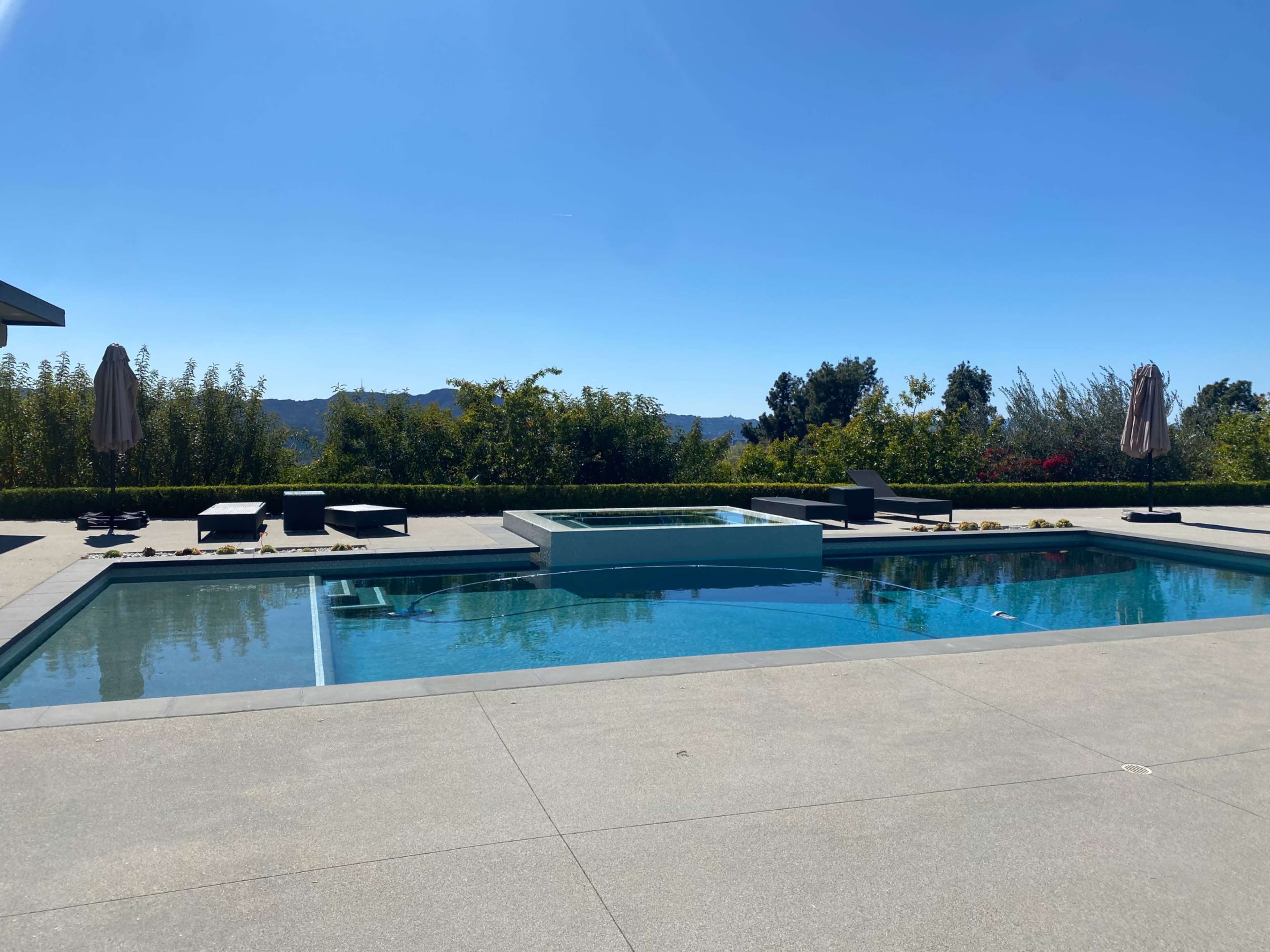 The image shows a modern outdoor pool area with a hot tub surrounded by lounge chairs and umbrellas, set against a backdrop of greenery and distant hills under a clear blue sky.