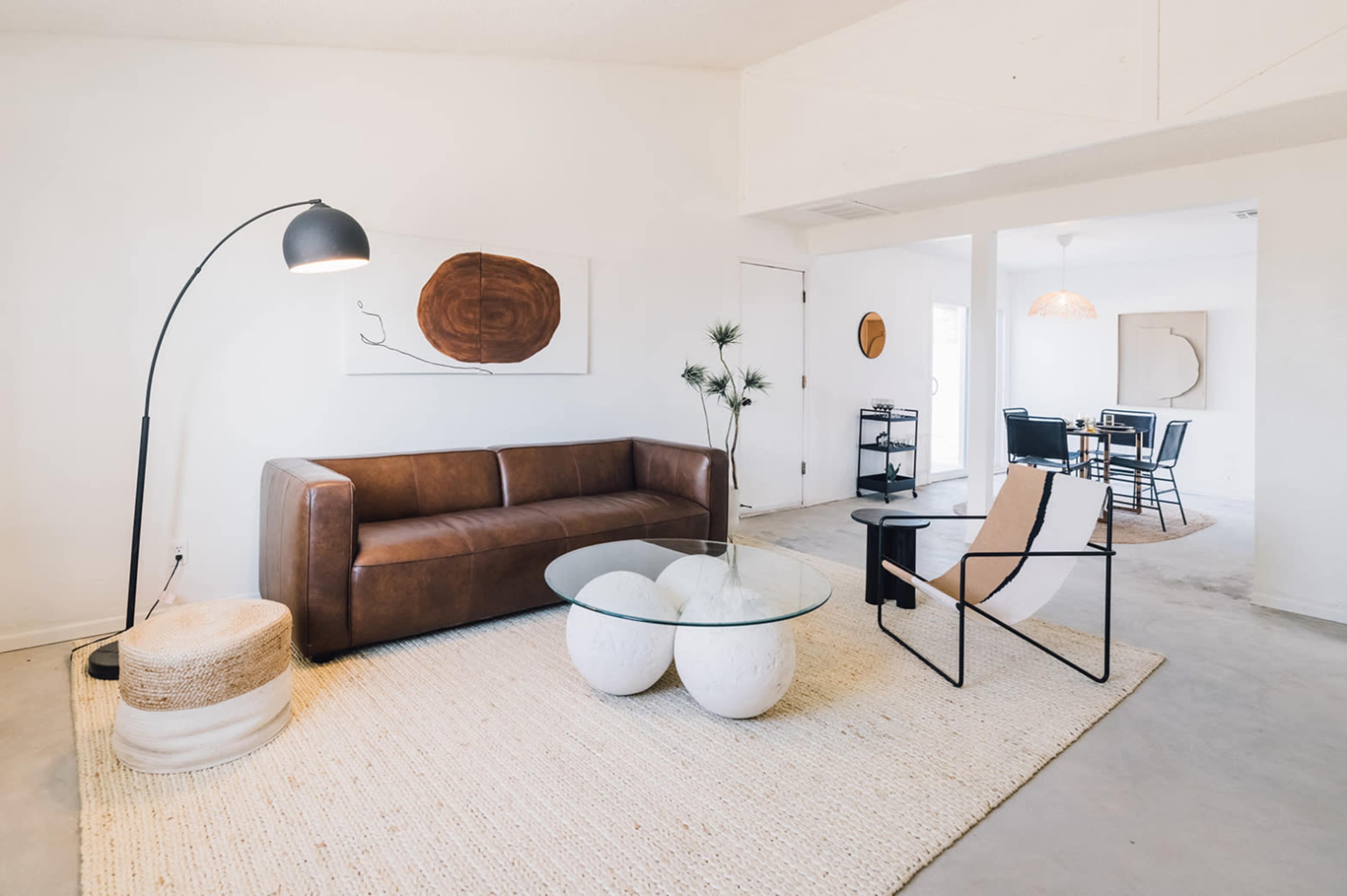 A contemporary living room features a brown leather sofa, a glass coffee table with rounded bases, and a floor lamp, set on a textured rug with an open layout leading to a dining area.