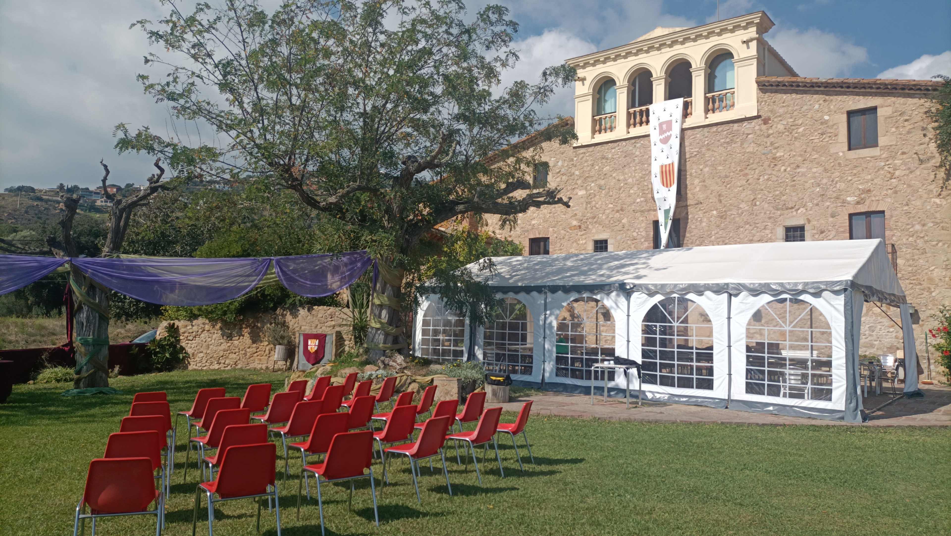 The image shows a garden setting with rows of red chairs arranged facing a white tent and a stone building in the background.