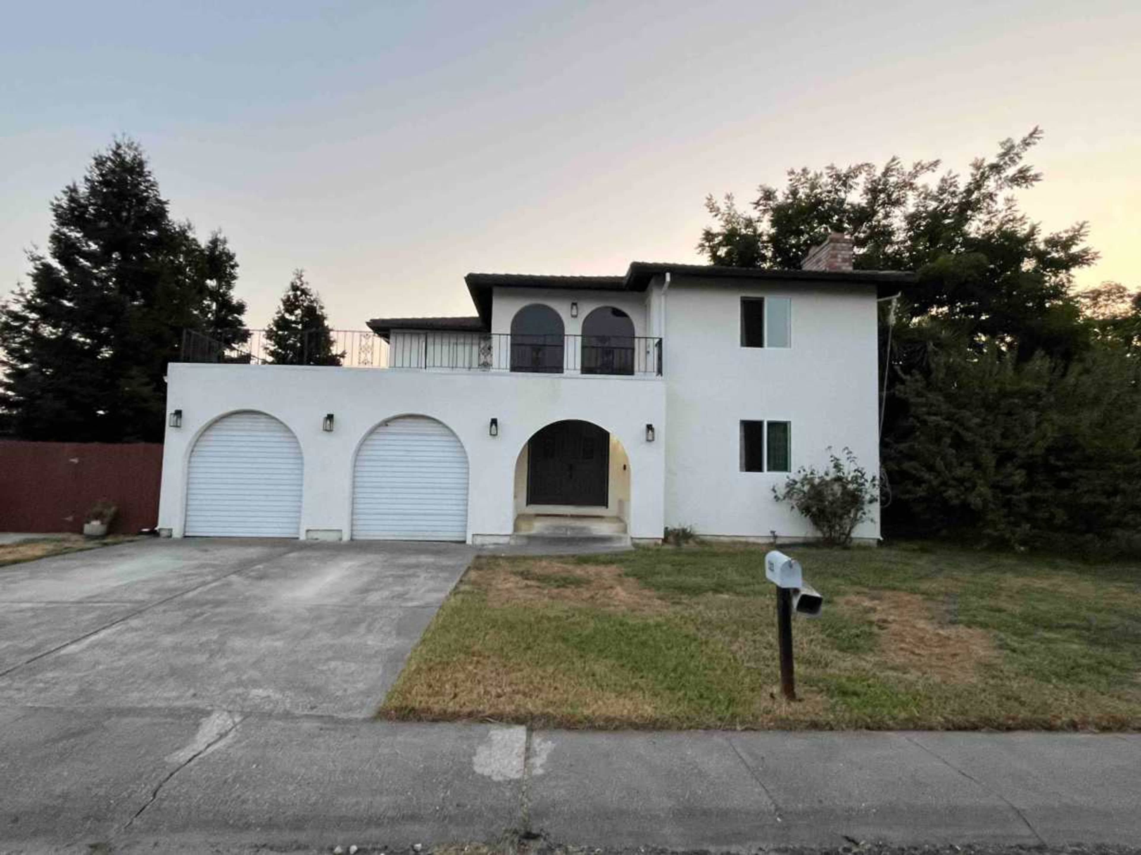 The image shows a two-story white house with a balcony, arched windows, and two garage doors, set on a grassy lot.