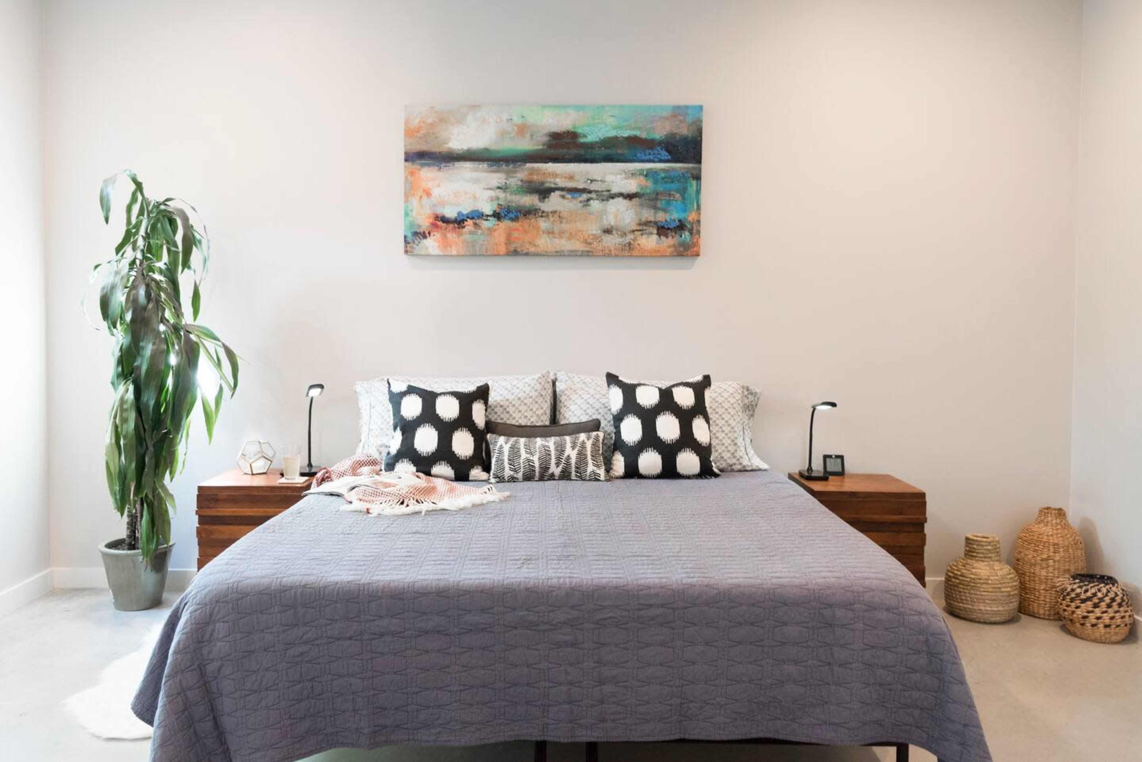 A neatly made bed with decorative pillows is positioned against a light-colored wall, accompanied by bedside tables and a tall plant in a pot.