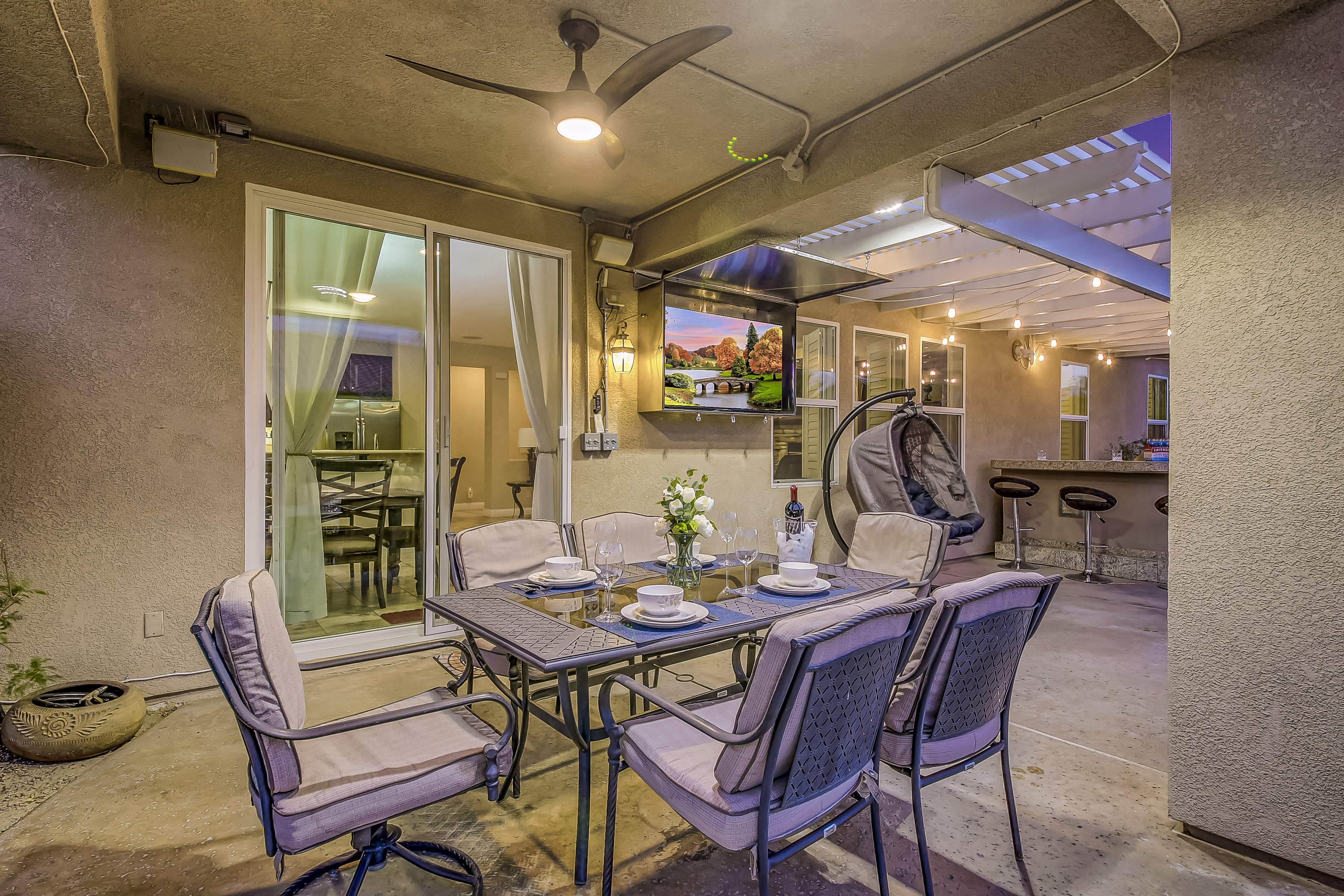 A patio area features a dining table with chairs, a bottle of wine, and a television, surrounded by a covered seating space.