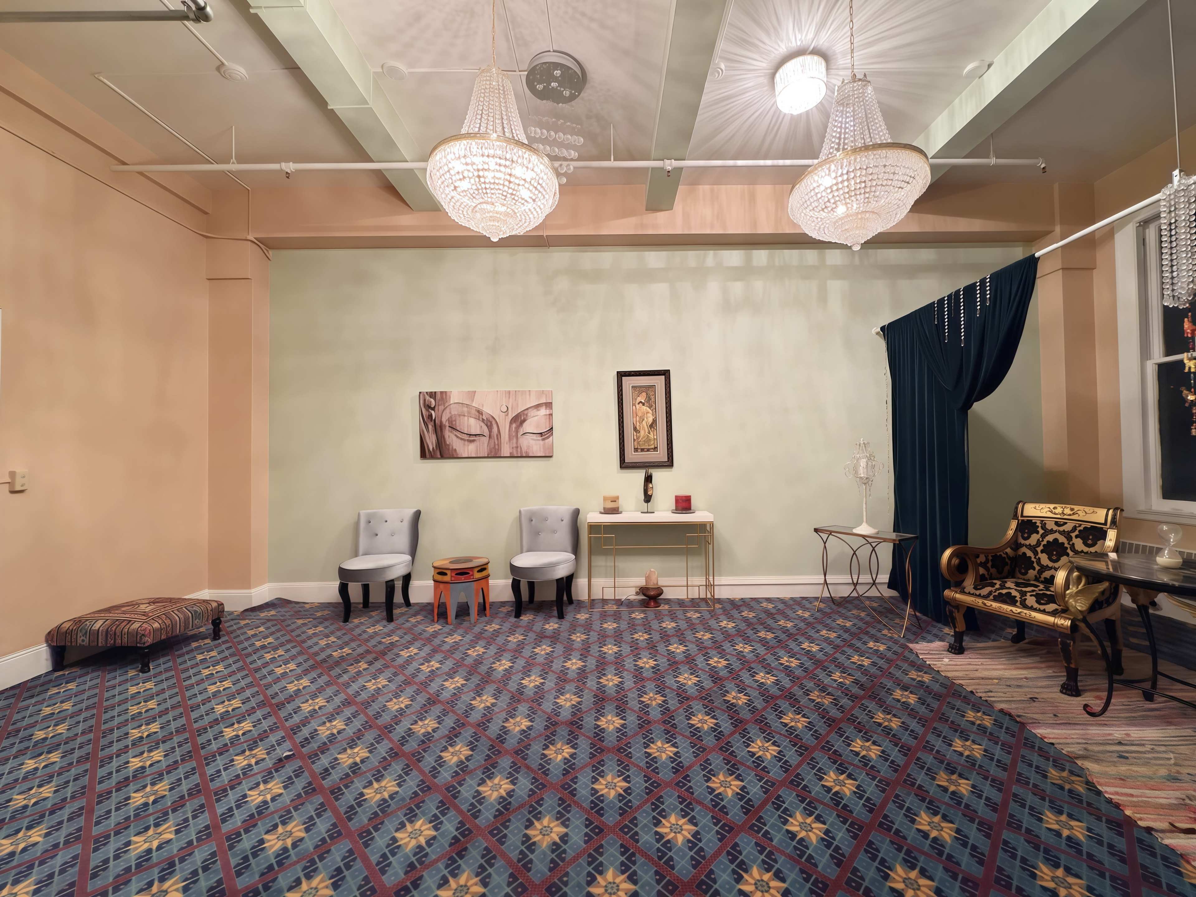 The room features ornate chandeliers, two chairs, a patterned rug, and a backdrop with decorative elements and artwork on the walls.