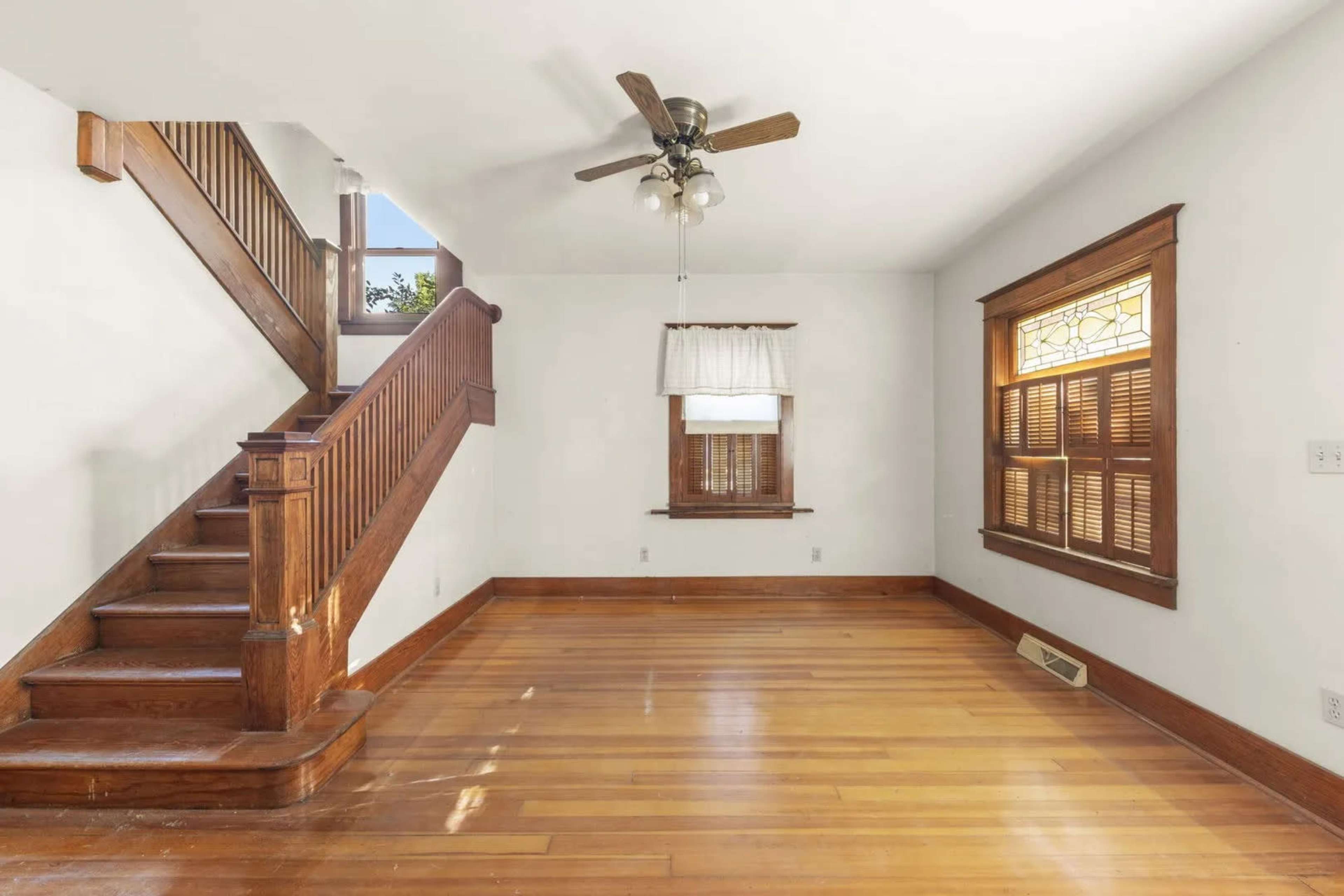 The image shows a spacious living room with hardwood floors, a staircase on the left, and a window featuring wooden blinds.