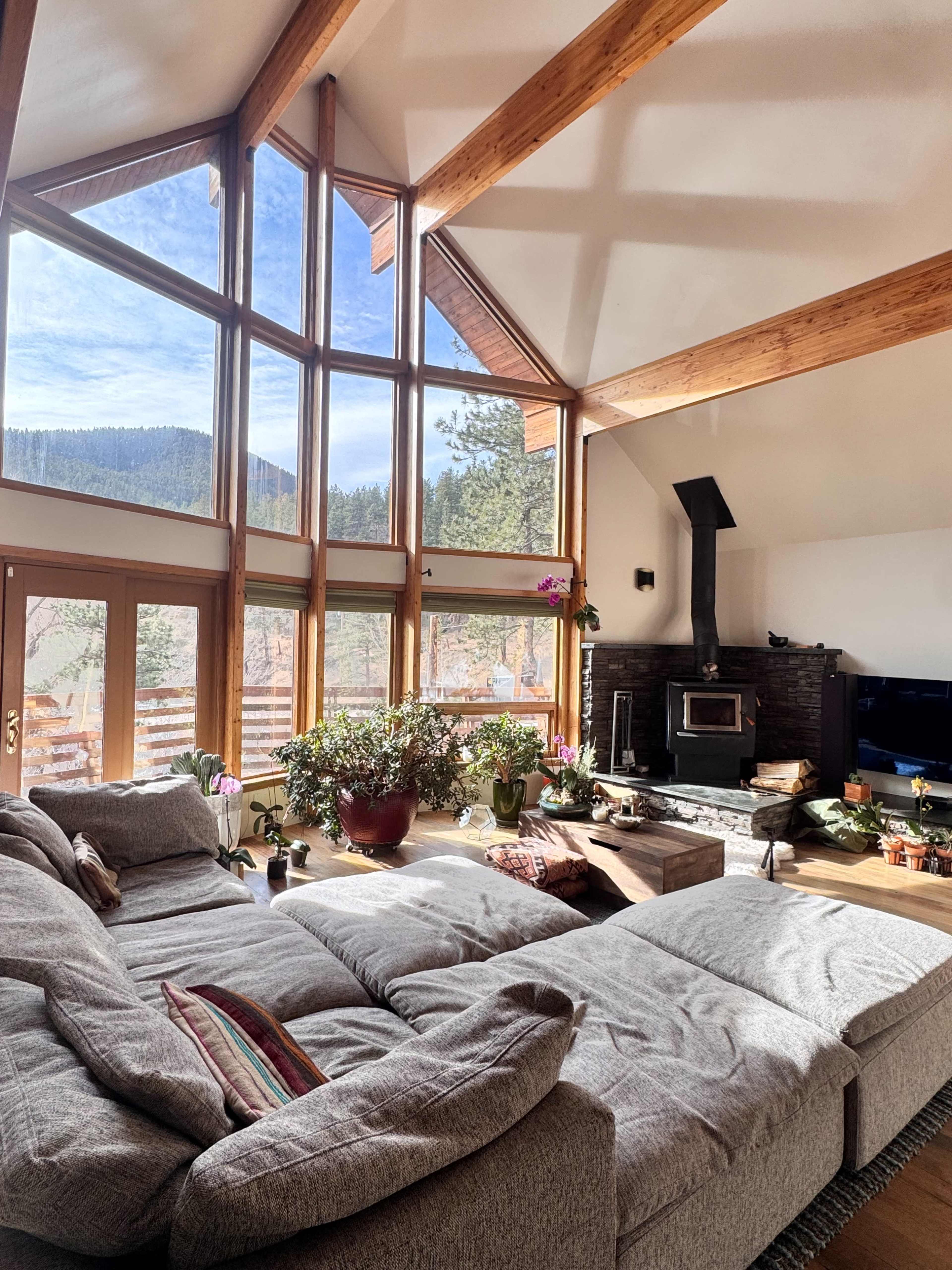 The image shows a spacious living room with large windows that provide a view of the mountains, featuring a cozy sectional sofa, a fireplace, and several plants on the coffee table.