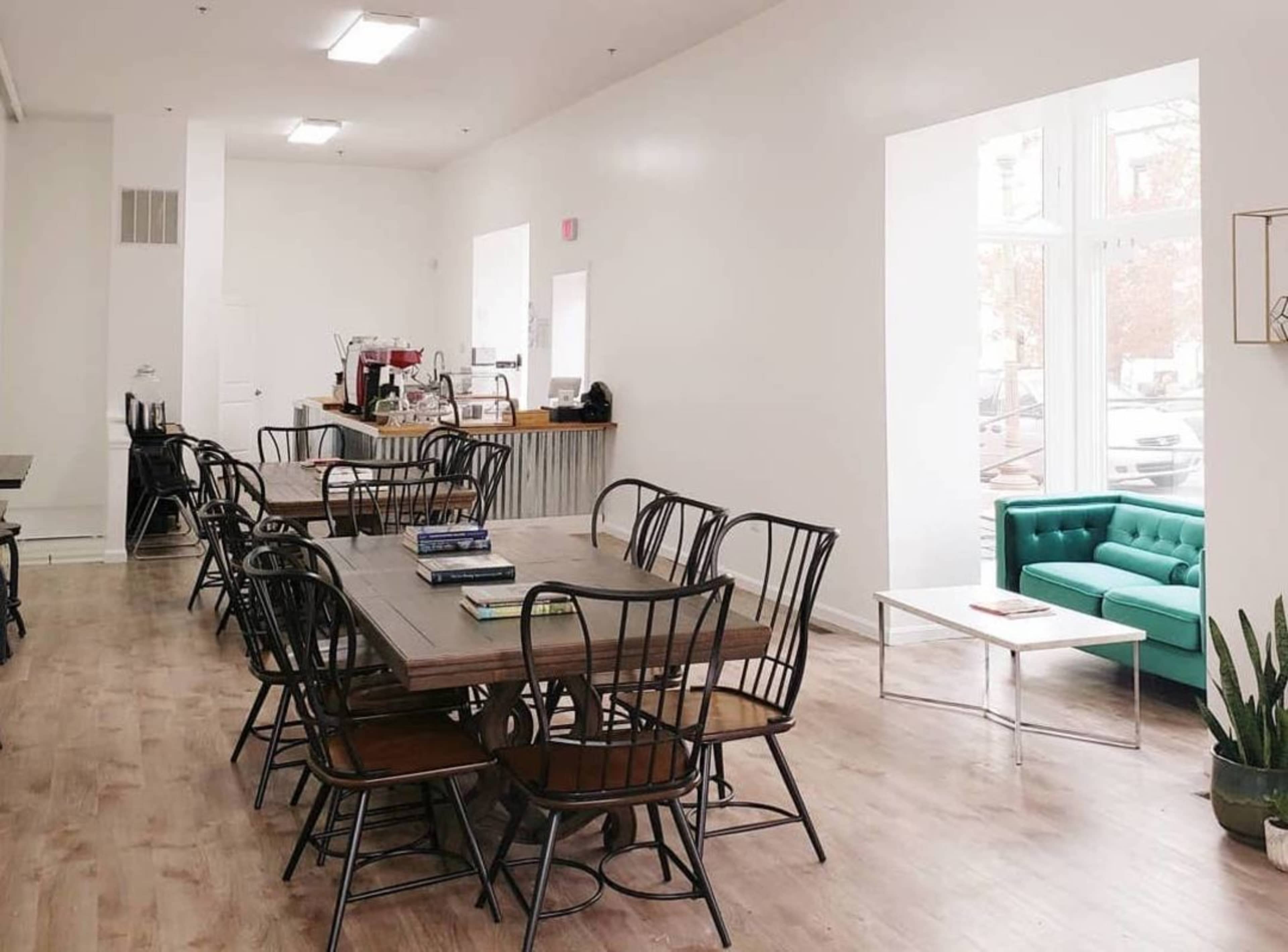 A bright, spacious café with wooden tables, black chairs, and a green couch along the wall.