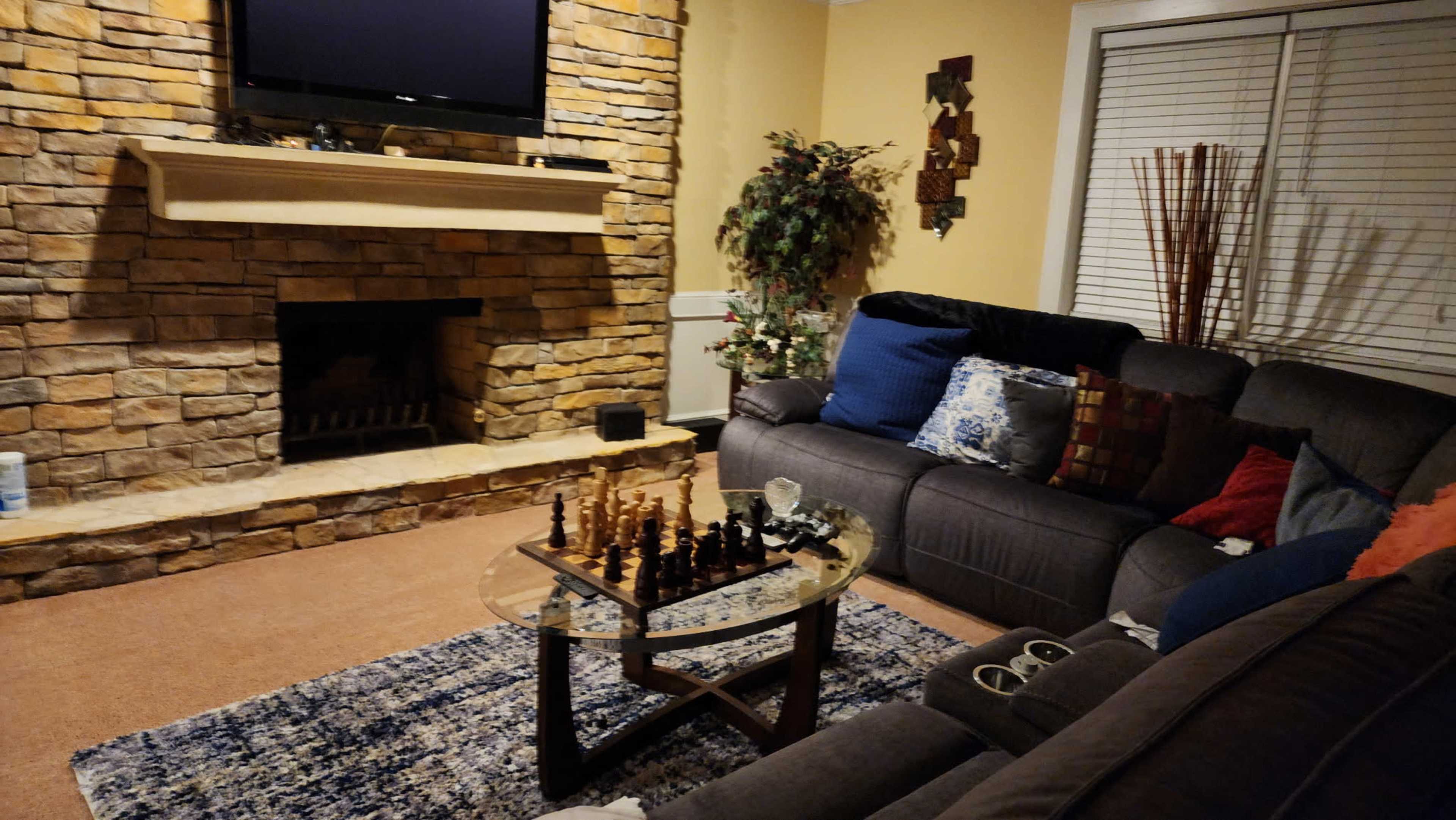 The image shows a cozy living room with a stone fireplace, a large sectional sofa, a glass coffee table containing a chess set, and a television mounted on the wall.