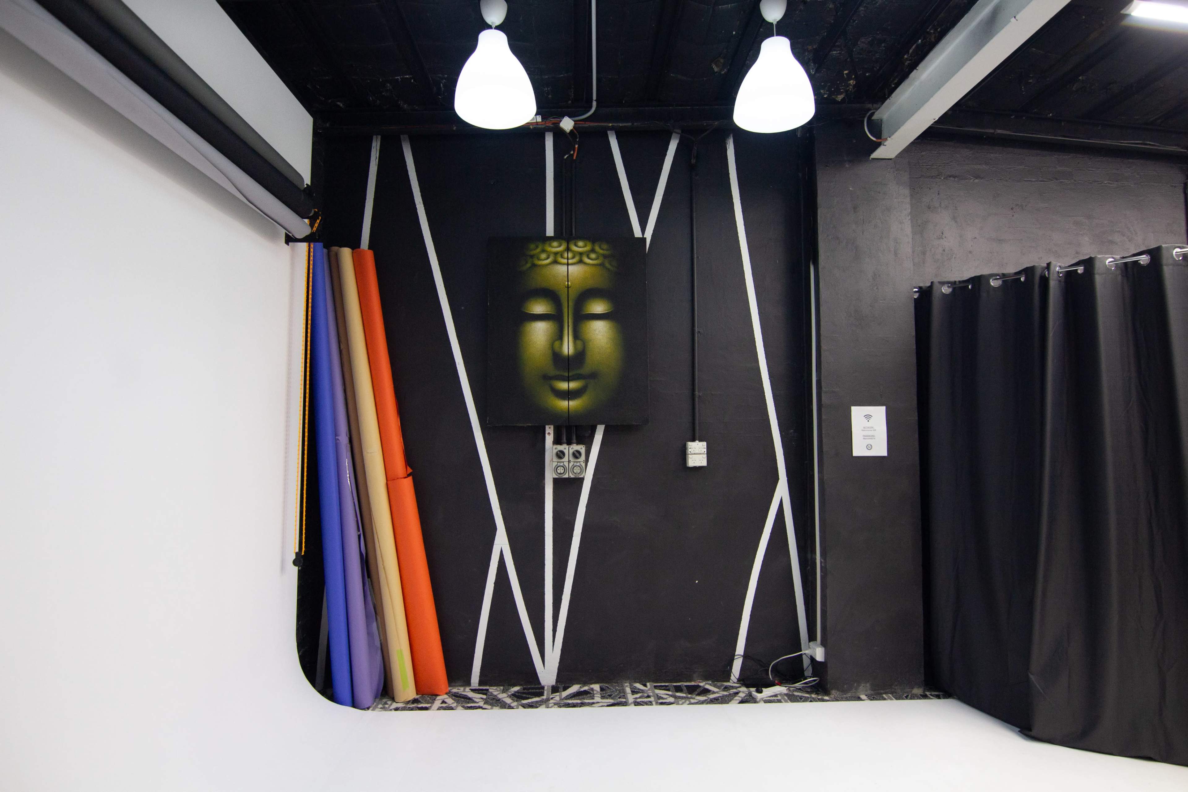 A photography studio interior with a black wall featuring a Buddha painting, colorful fabric rolls, and black curtains.