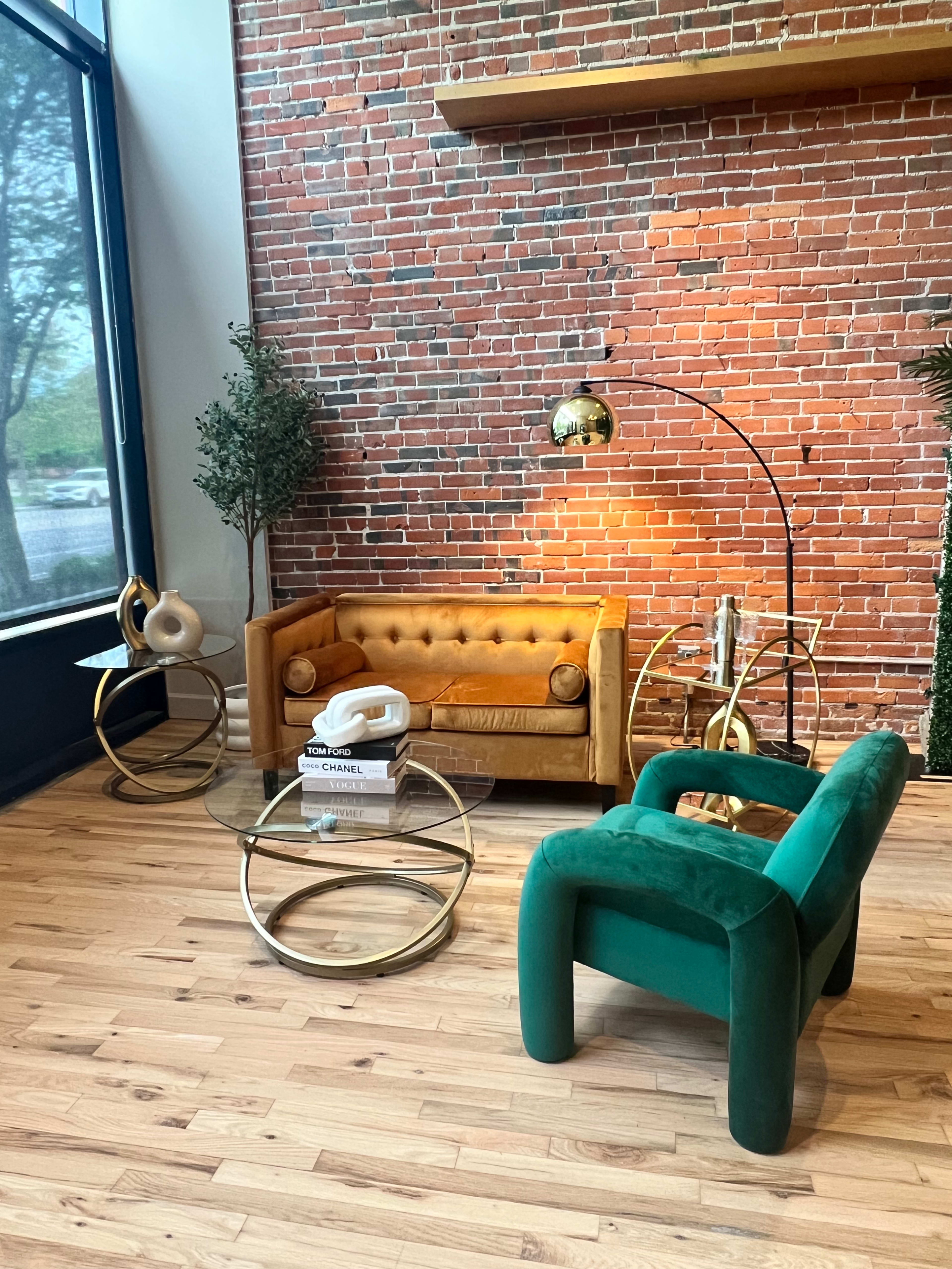 A stylish seating area featuring a vintage yellow sofa, a green armchair, a glass coffee table, and a modern floor lamp against a brick wall.
