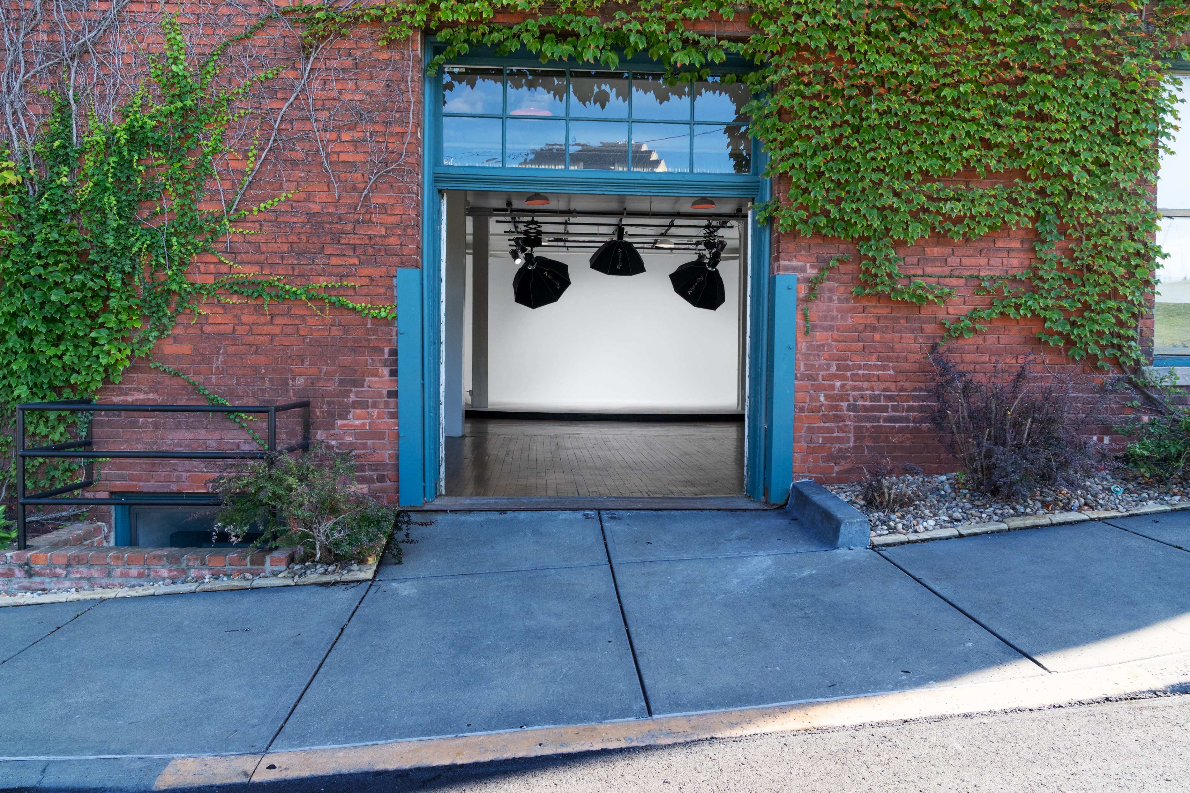 The image shows an open entrance to a brick building with ivy climbing the walls, revealing a spacious interior with studio lights hanging from the ceiling.