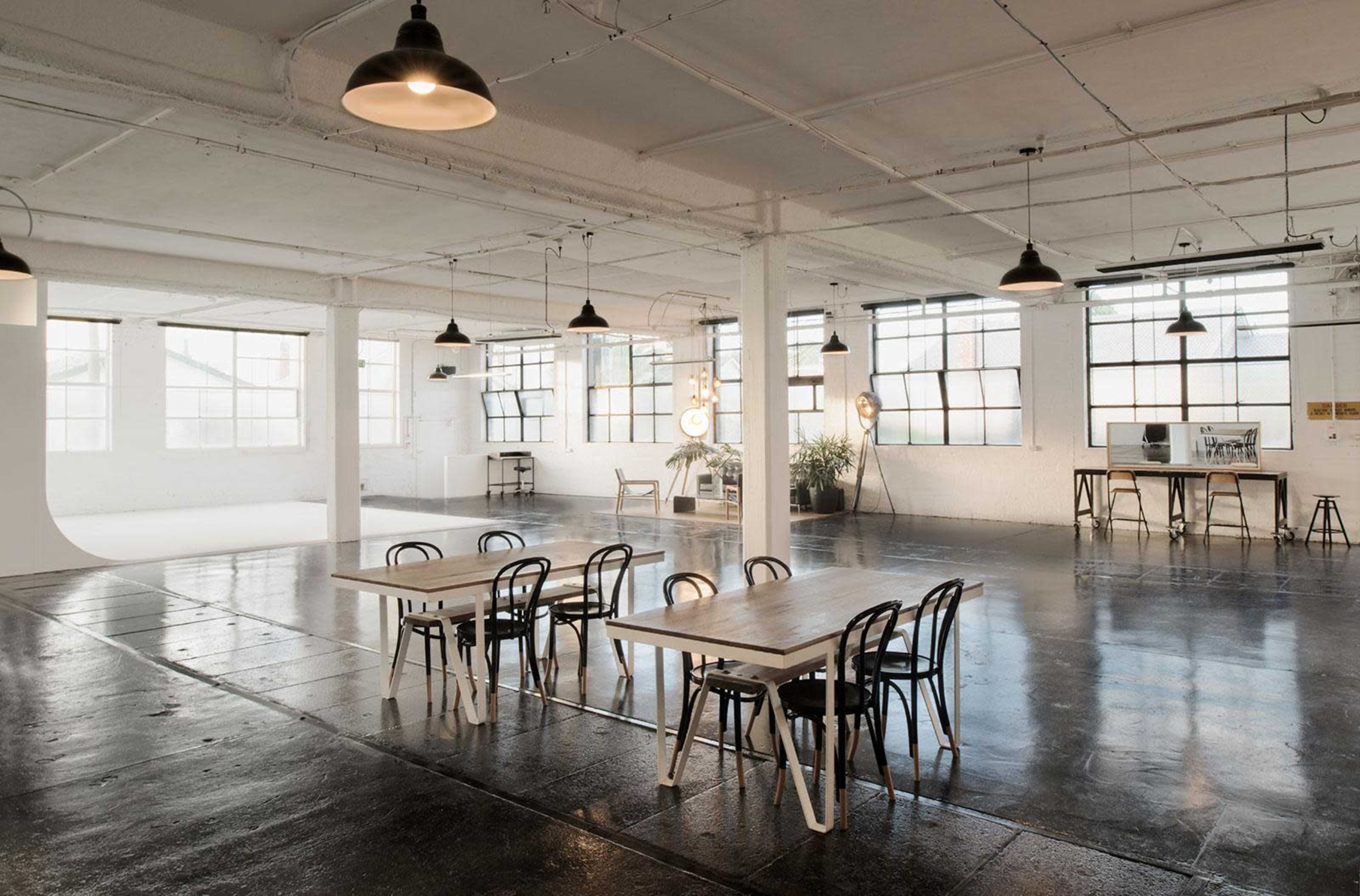 An industrial-style room with polished concrete floors, large windows, and several wooden tables and chairs arranged for a creative workspace.