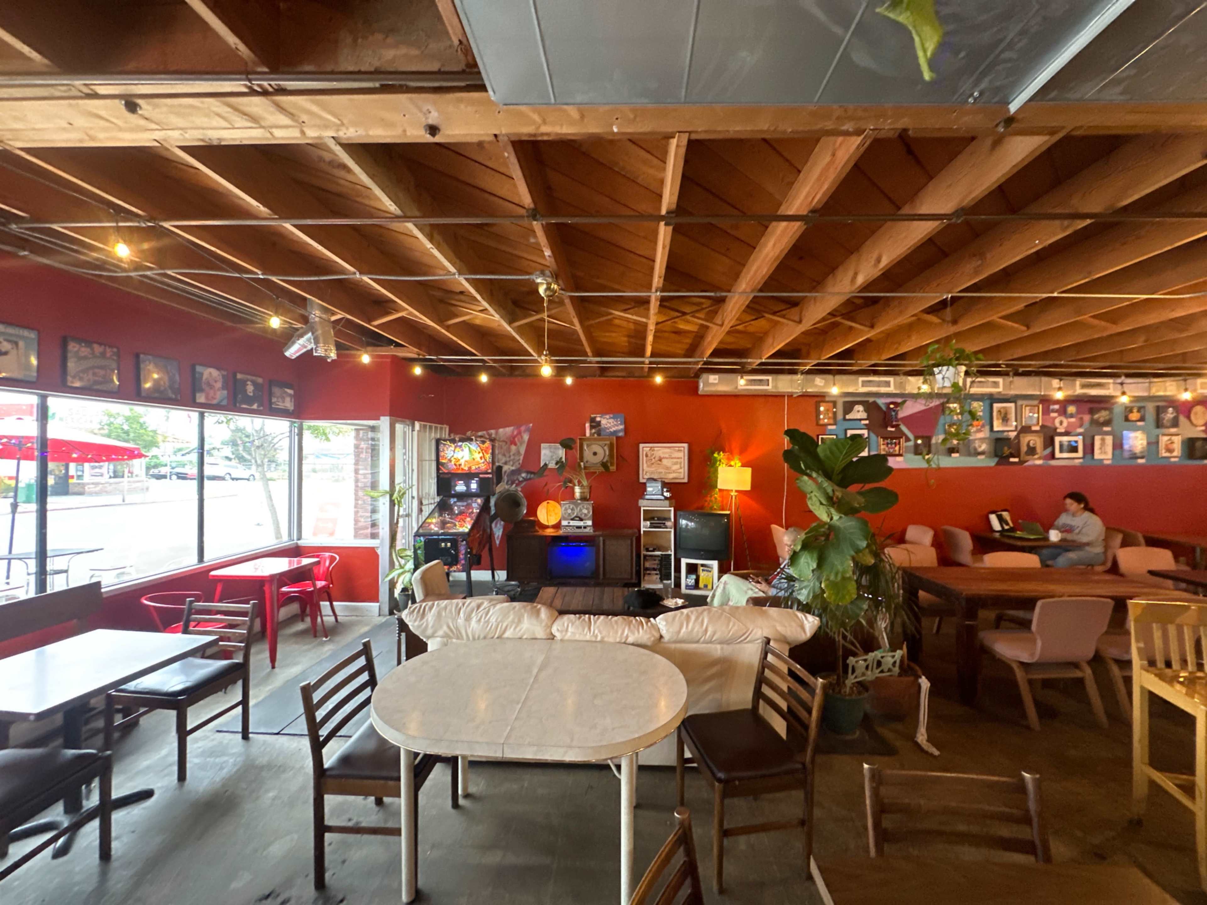 The interior of a casual café features various seating arrangements, a mix of tables and chairs, and a few arcade games against a red wall adorned with photographs.