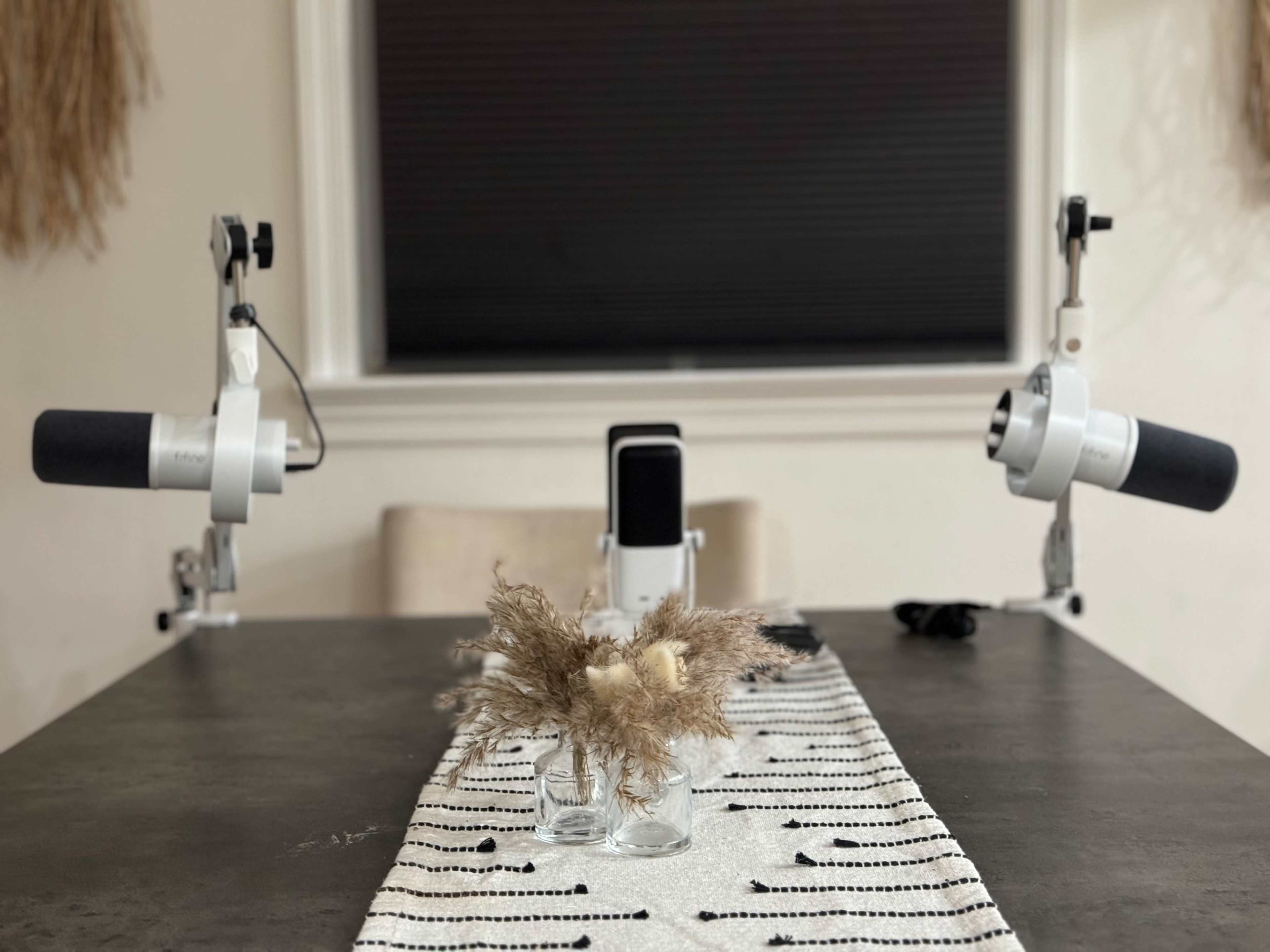 A table setting features two microphones on adjustable stands facing a central arrangement of dried plants in a small glass vase.