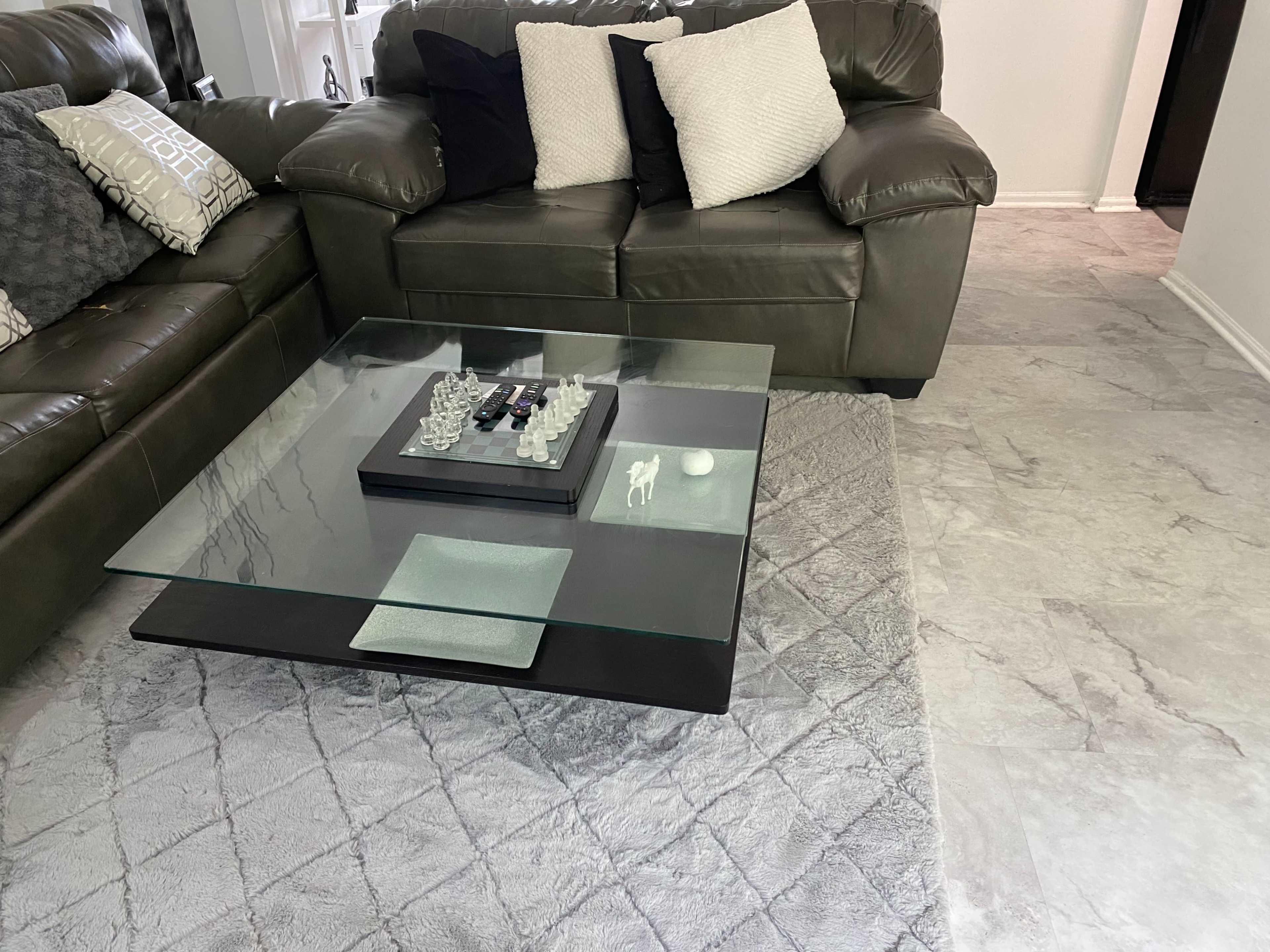 A living room features a green leather couch with decorative pillows, a glass coffee table holding a chess set and a decorative tray, and a light-colored rug on a tiled floor.