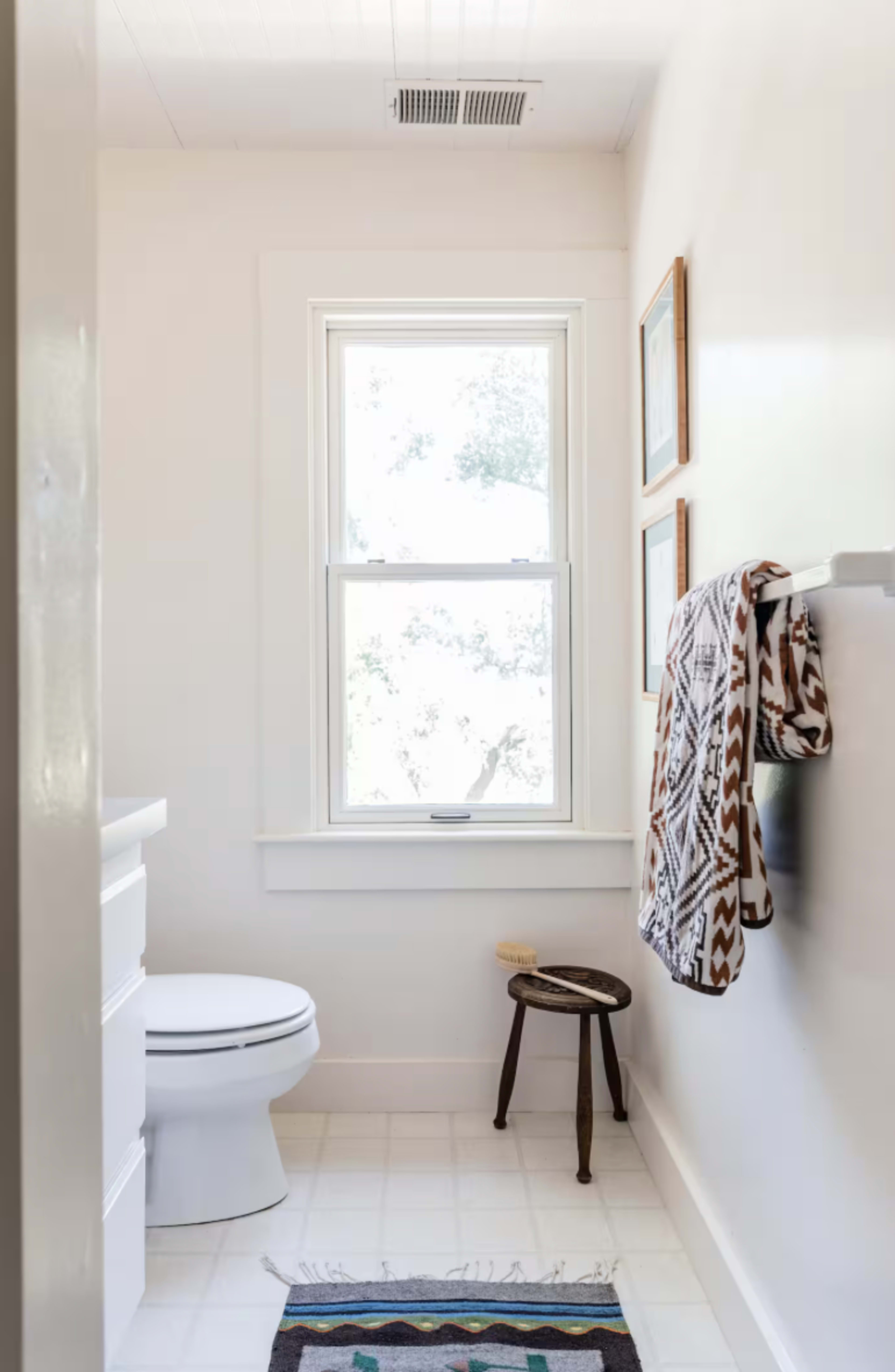 The image features a bright, minimalist bathroom with a white toilet, a small stool, and a window allowing natural light in.