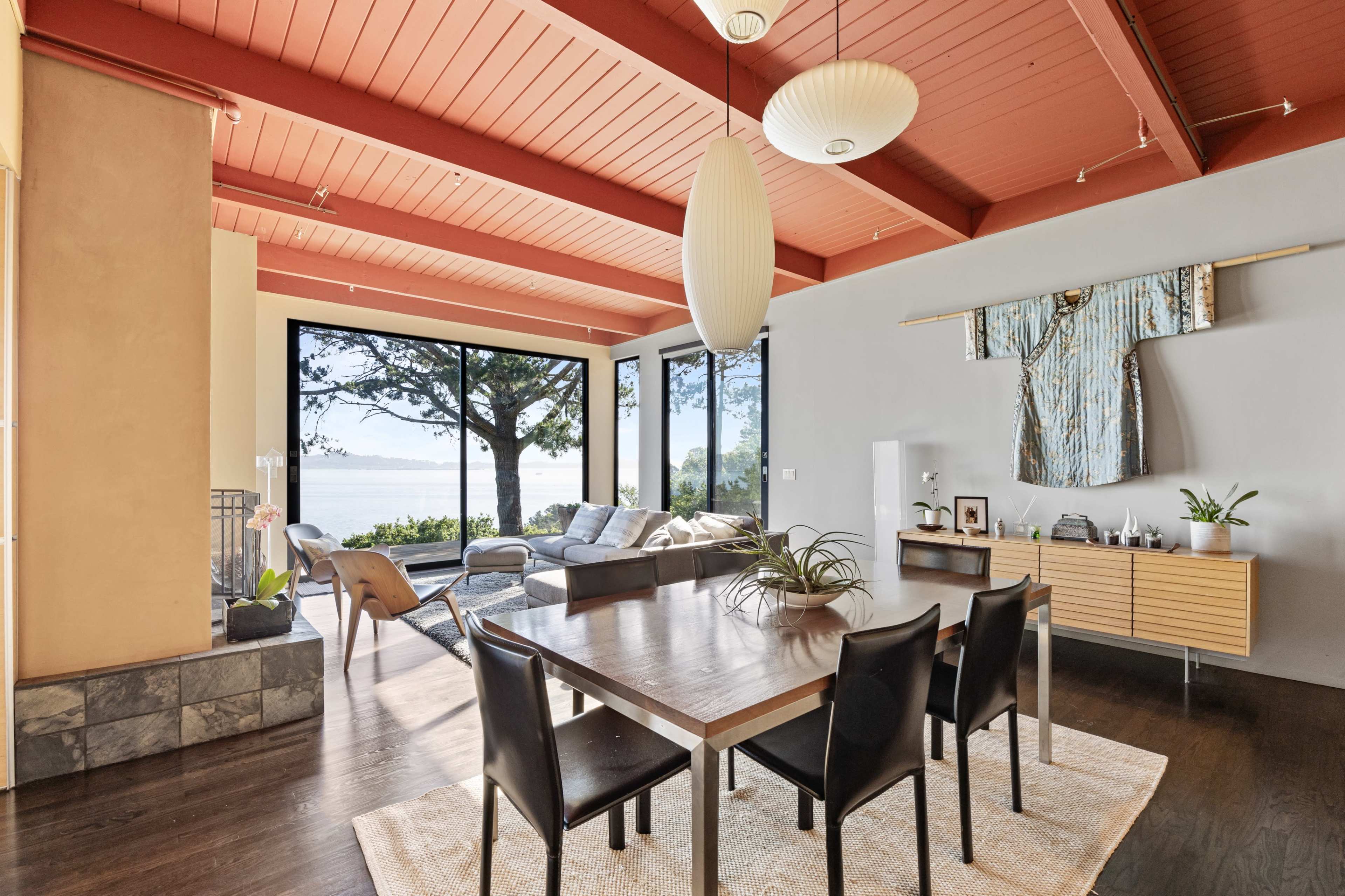 A modern interior features a dining area with a wooden table and black chairs, overlooking large windows that reveal a view of a body of water and trees outside.