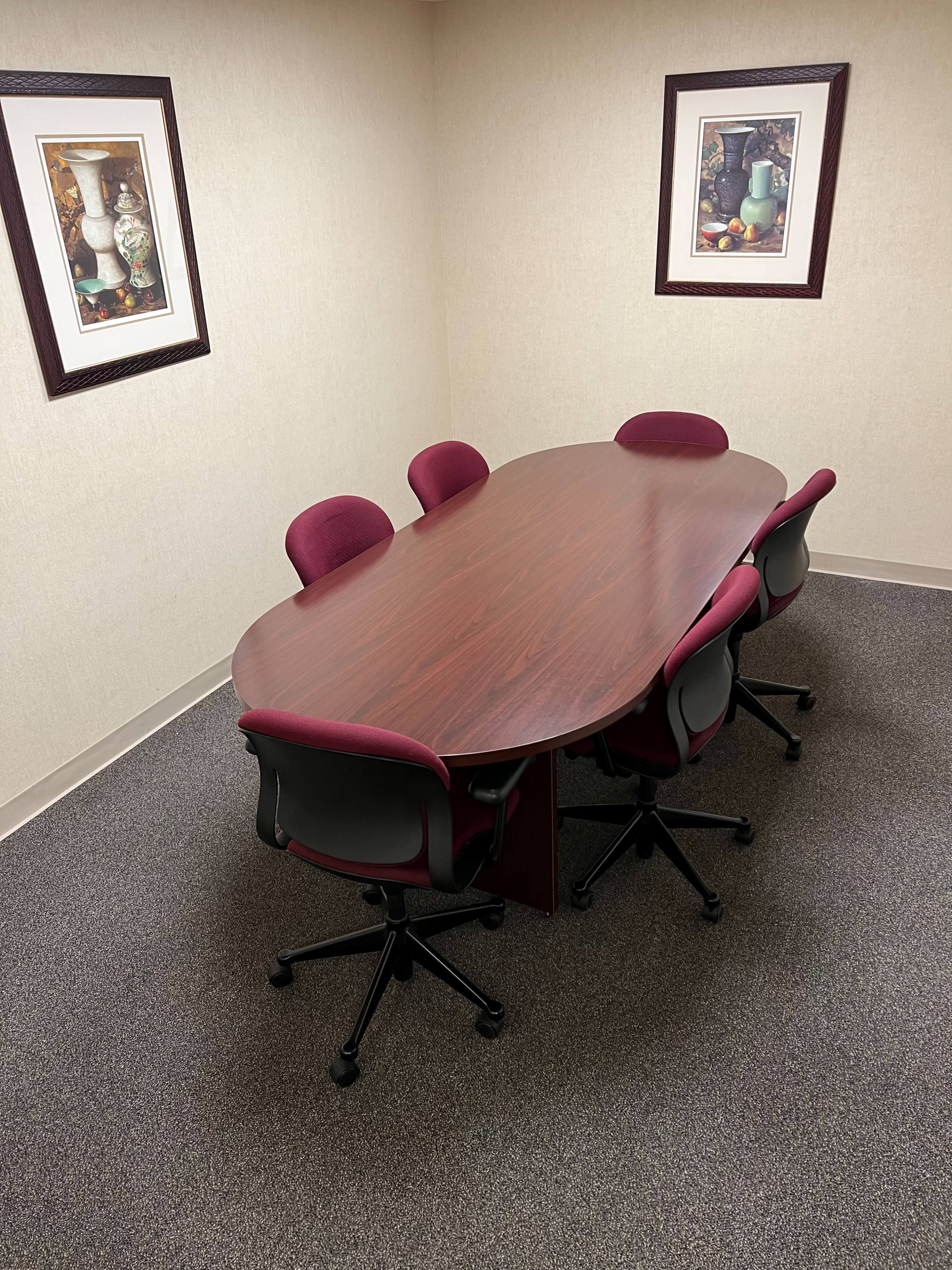A wooden oval conference table with five burgundy chairs is placed in a small, carpeted meeting room adorned with two framed artwork pieces on the walls.