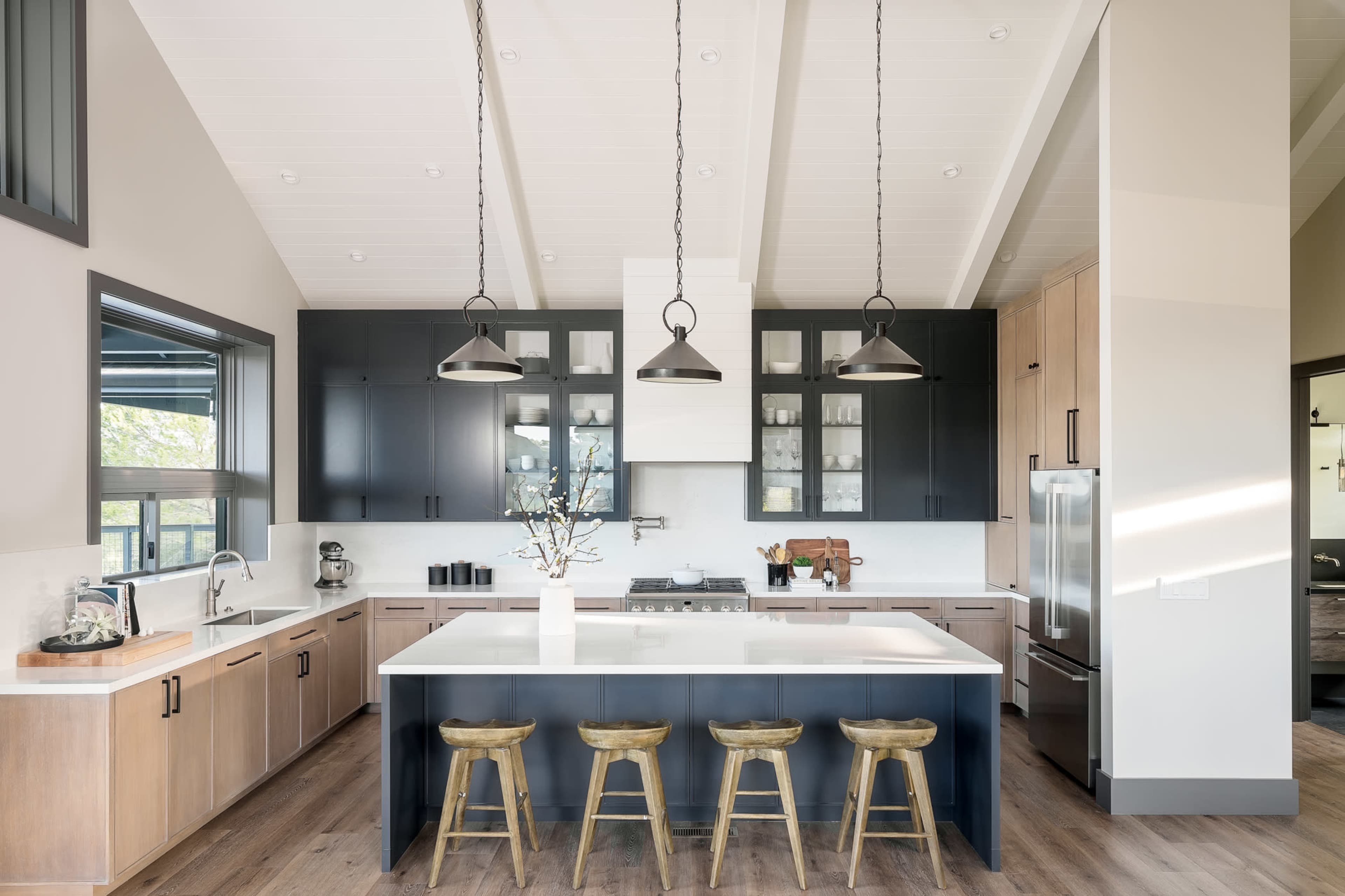 A modern kitchen featuring a large island with barstools, dark cabinetry, and a light countertop, complemented by pendant lighting and wooden flooring.