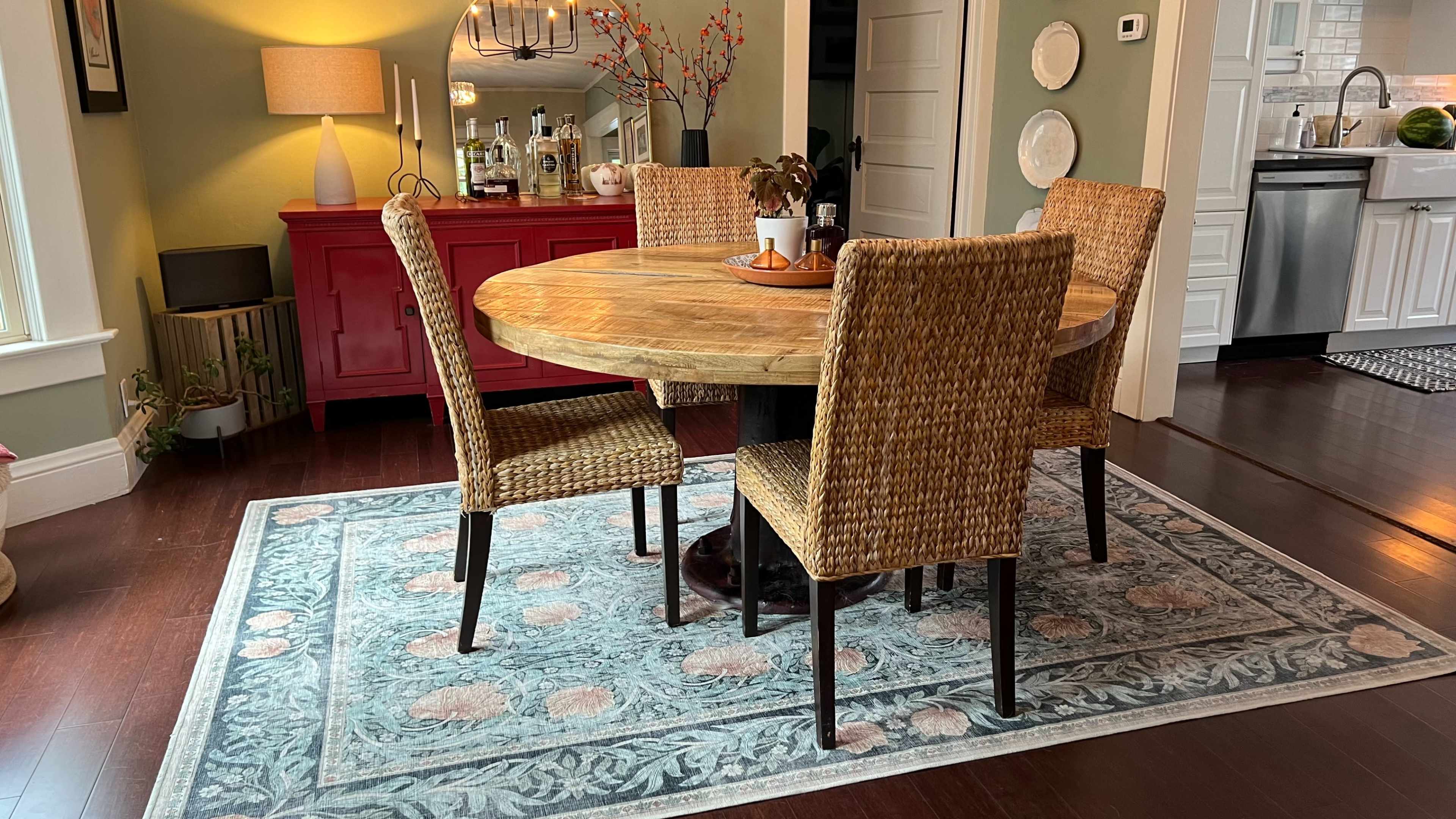 A round wooden dining table with four woven chairs is set on a patterned rug in a well-lit dining area.
