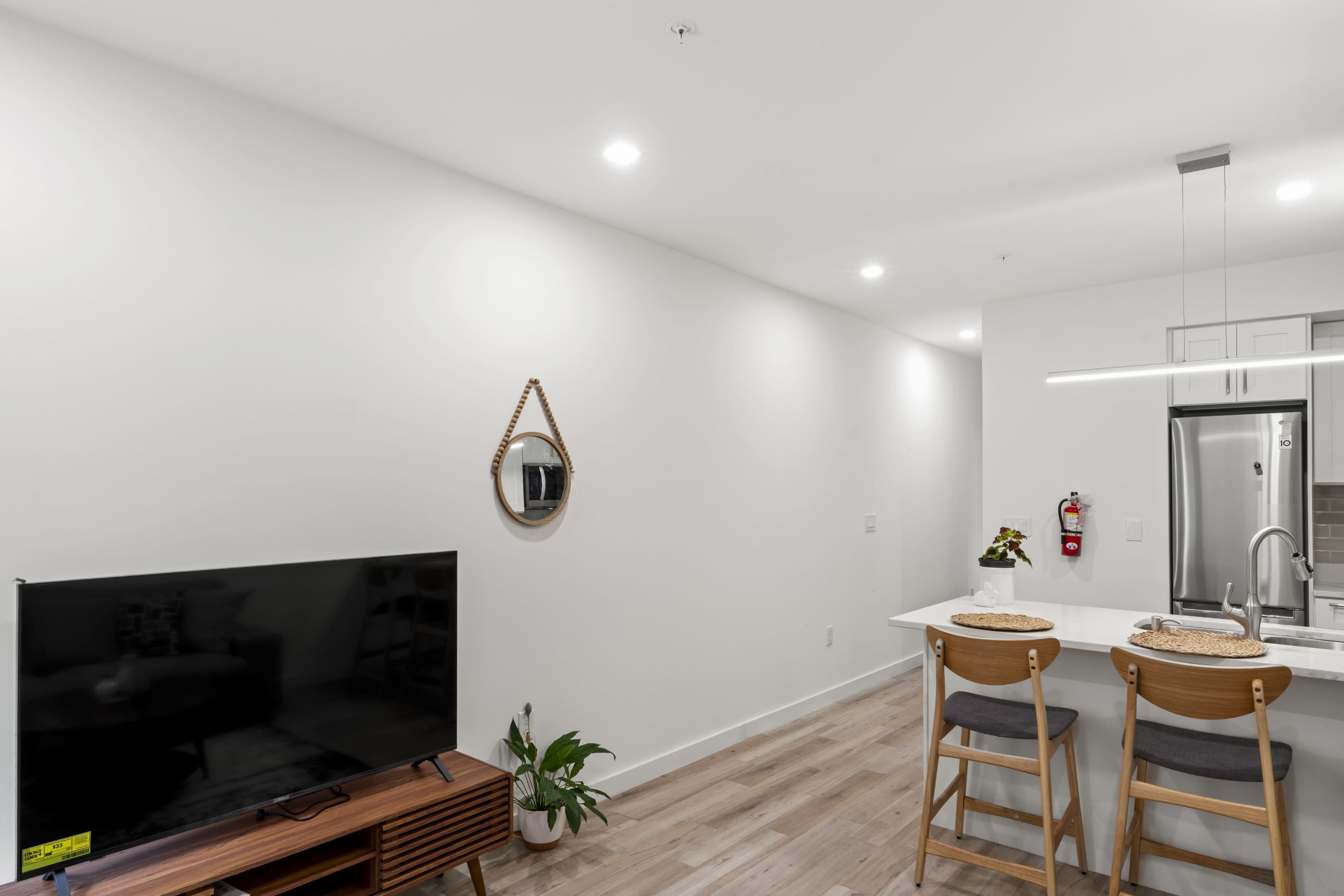 The image shows a modern living space with a television, a kitchen island with two bar stools, and a minimalistic aesthetic featuring light walls and wooden flooring.