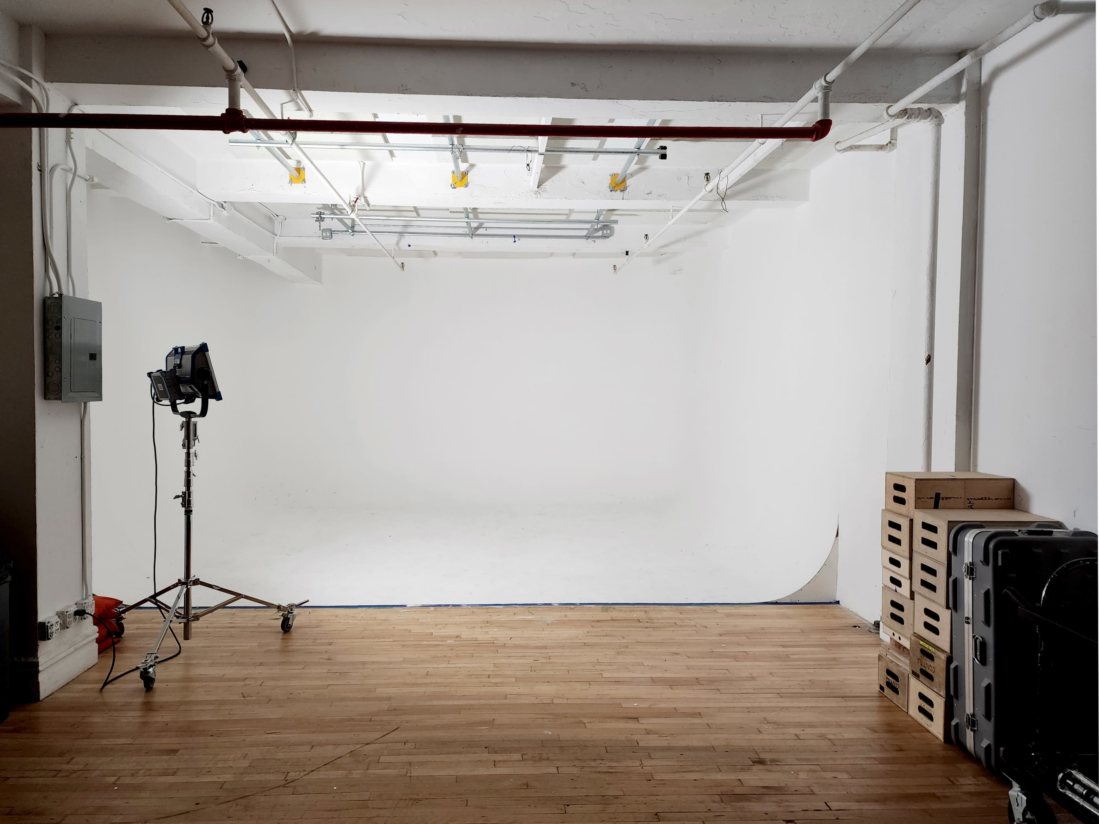 The image depicts a spacious, empty photography studio with white walls, hardwood flooring, and equipment including a light stand and storage boxes.
