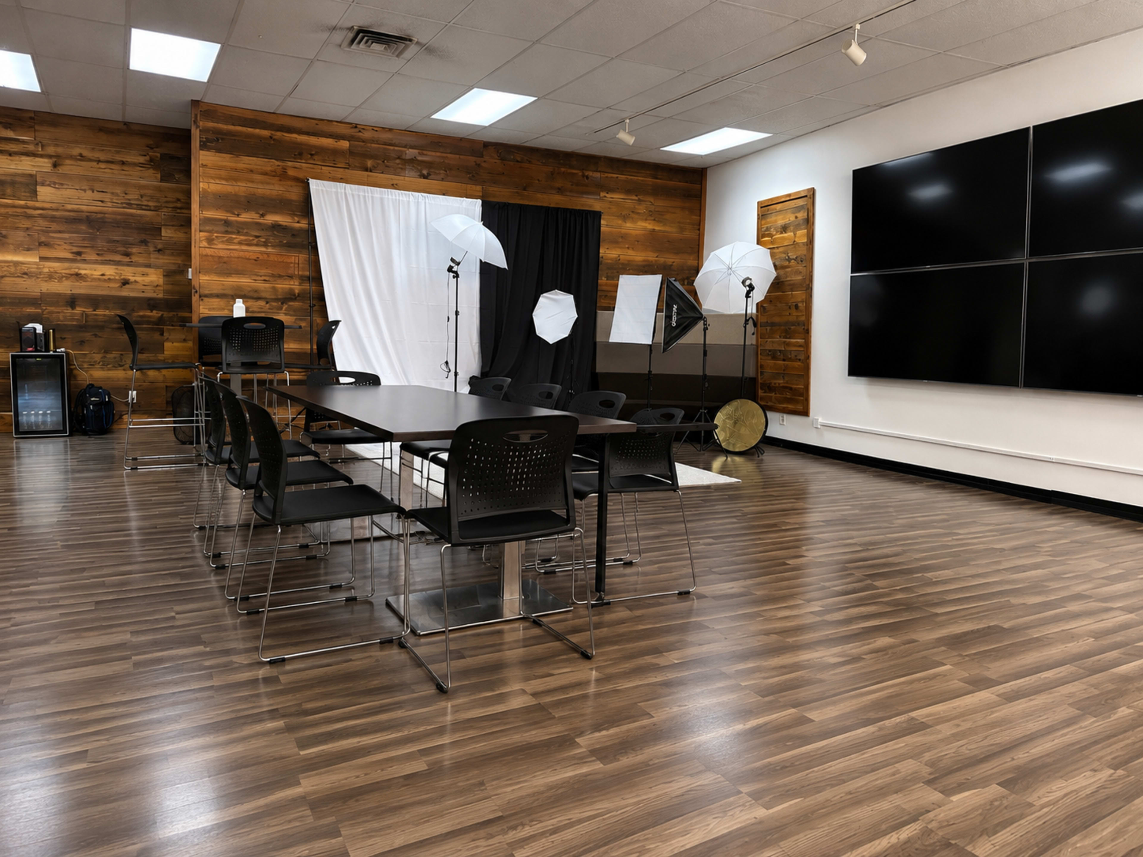 The image shows a modern conference room with a large table and black chairs, wooden walls, and photography equipment set up in the background.