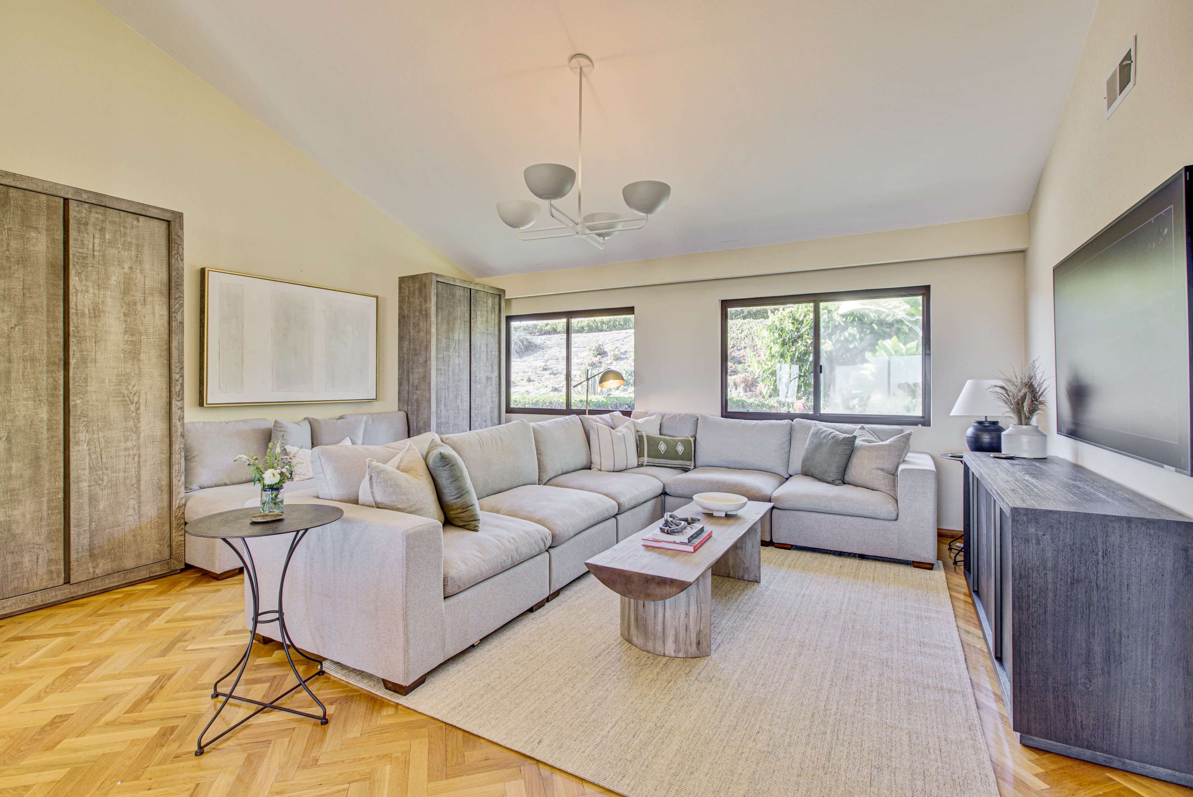 A spacious living room features a large sectional sofa, a circular side table, and a flat-screen TV, with large windows allowing natural light and views of greenery outside.