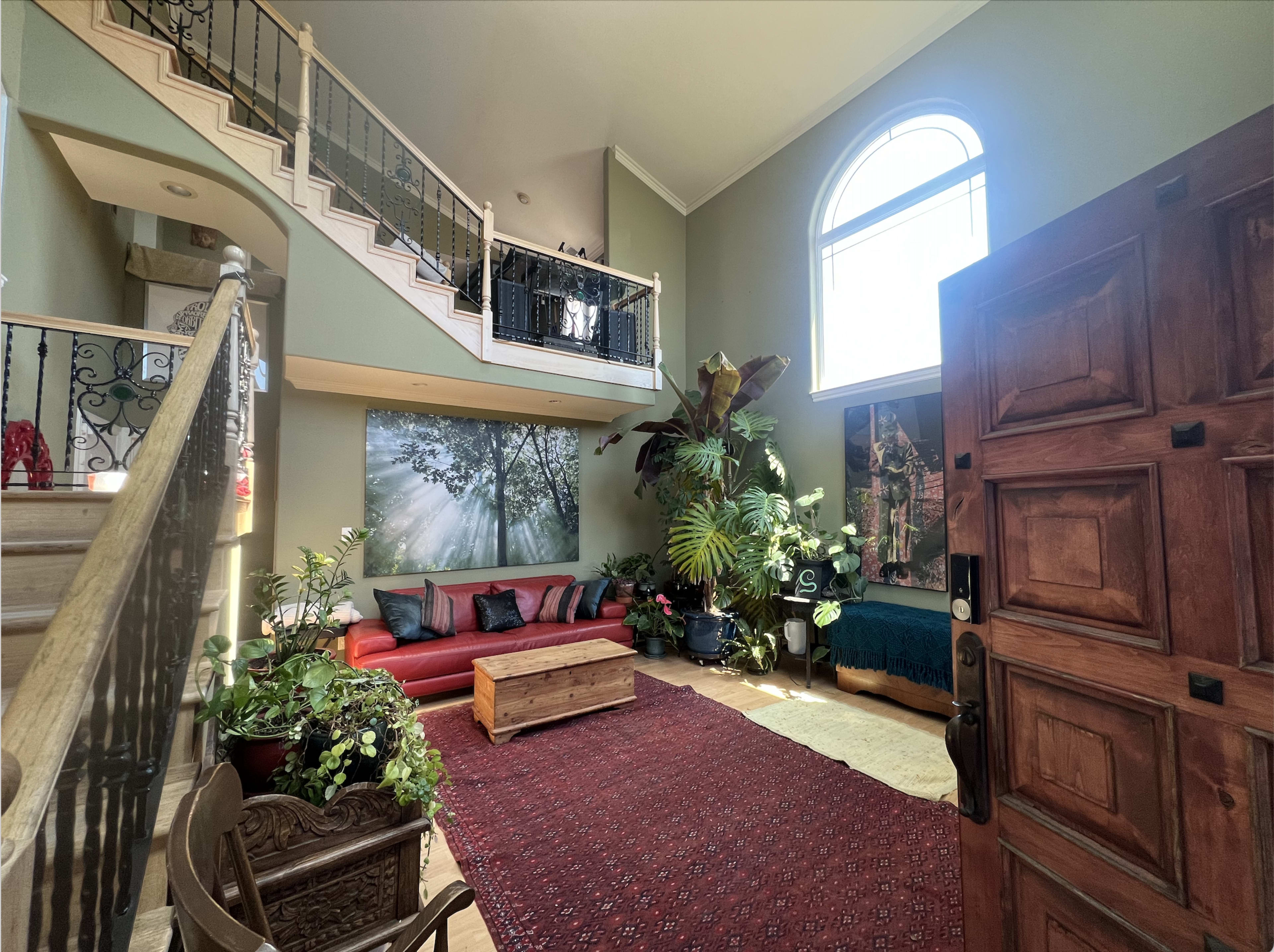 The image shows a spacious entryway with a wooden front door, a staircase leading to a second floor, and a red sofa surrounded by plants and a large wall mural.