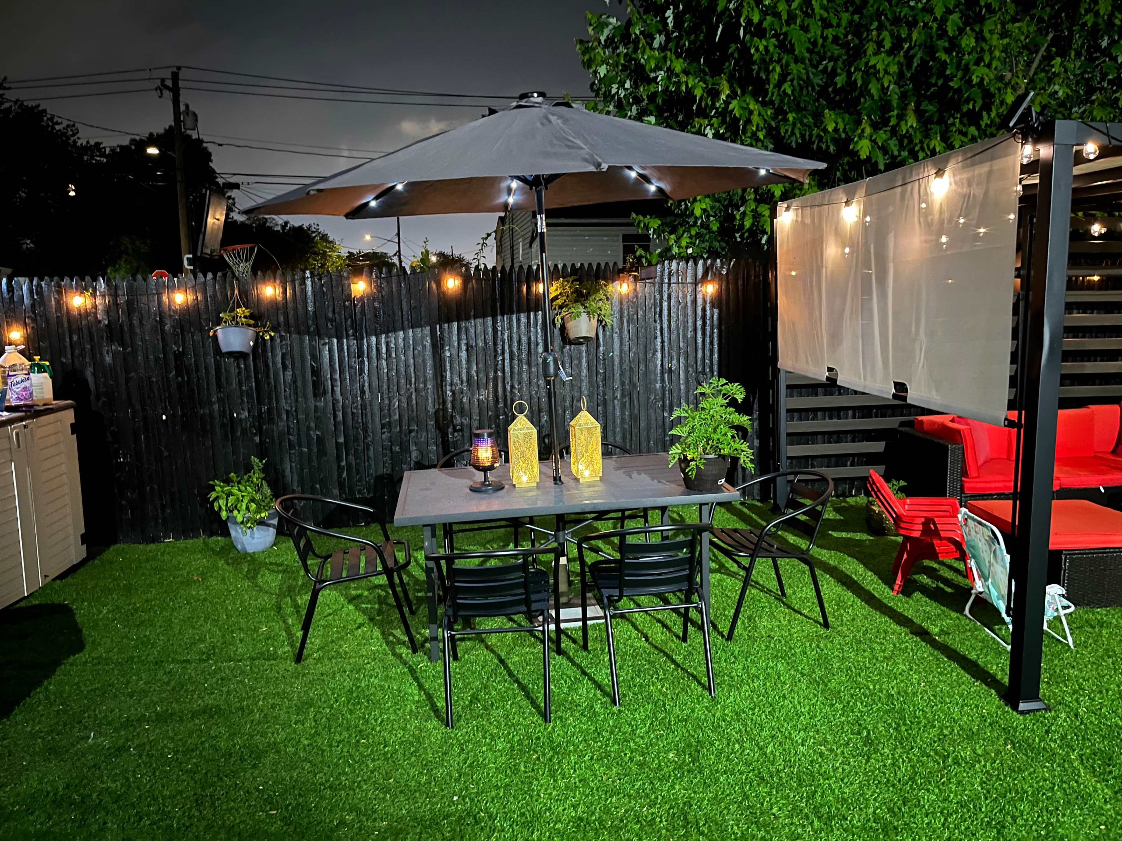 A patio features a dining table with chairs under an illuminated umbrella, surrounded by decorative plants and string lights against a dark sky.