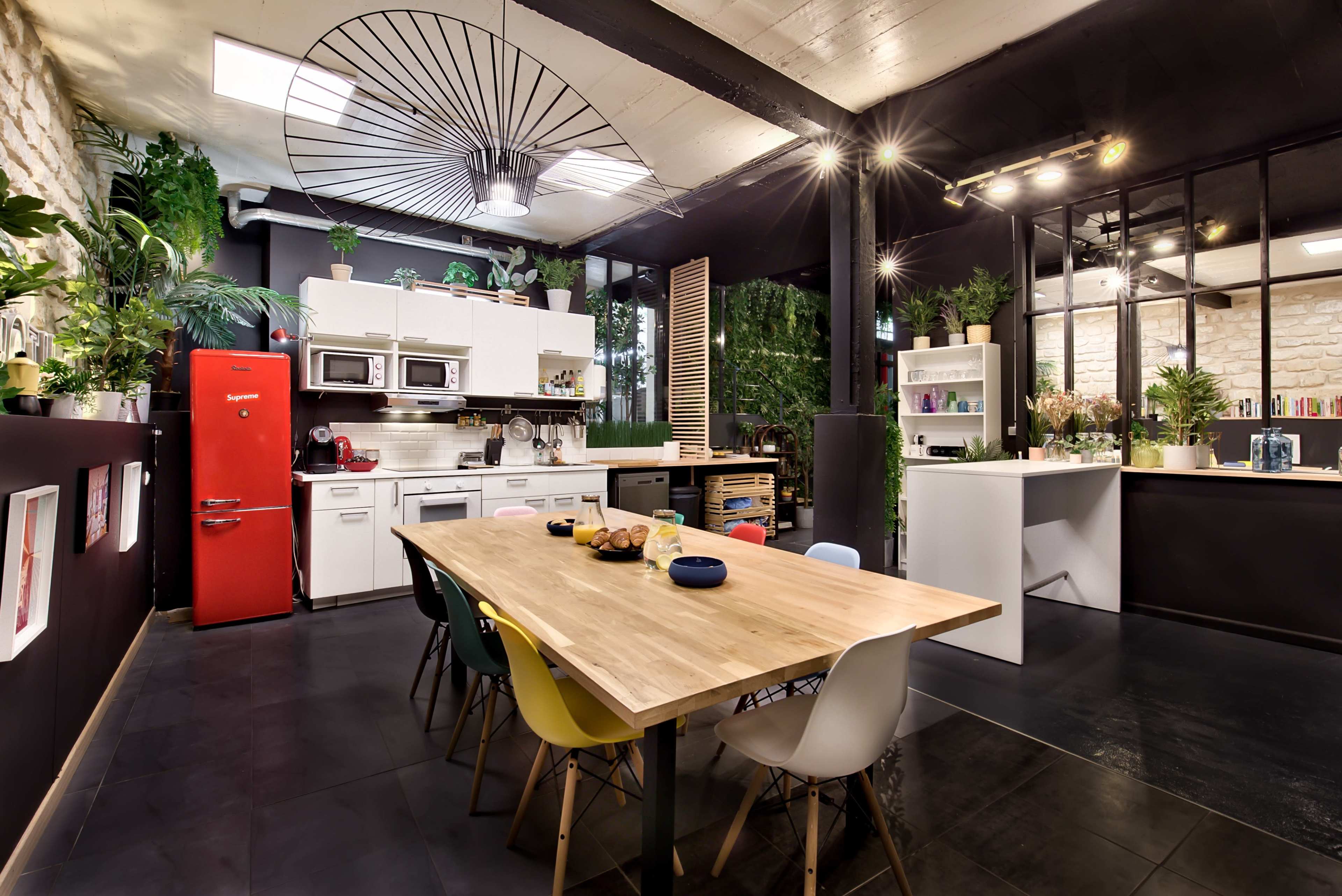 A modern kitchen and dining area features a large wooden table surrounded by colorful chairs, with plants and a bright red refrigerator accenting the space.