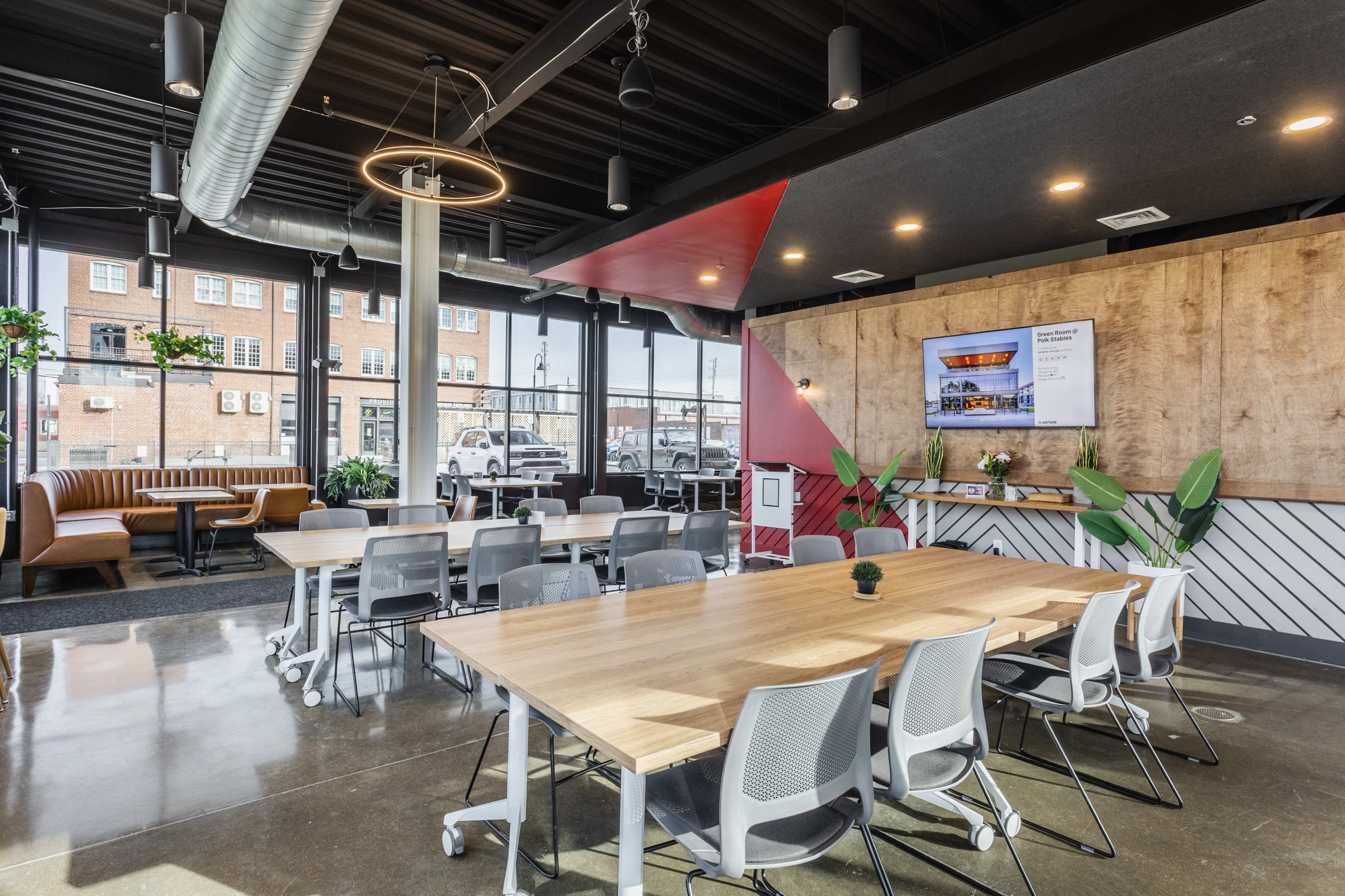 The image shows a modern conference room featuring a large wooden table with rolling chairs, a wall-mounted screen, and large windows that provide natural light.