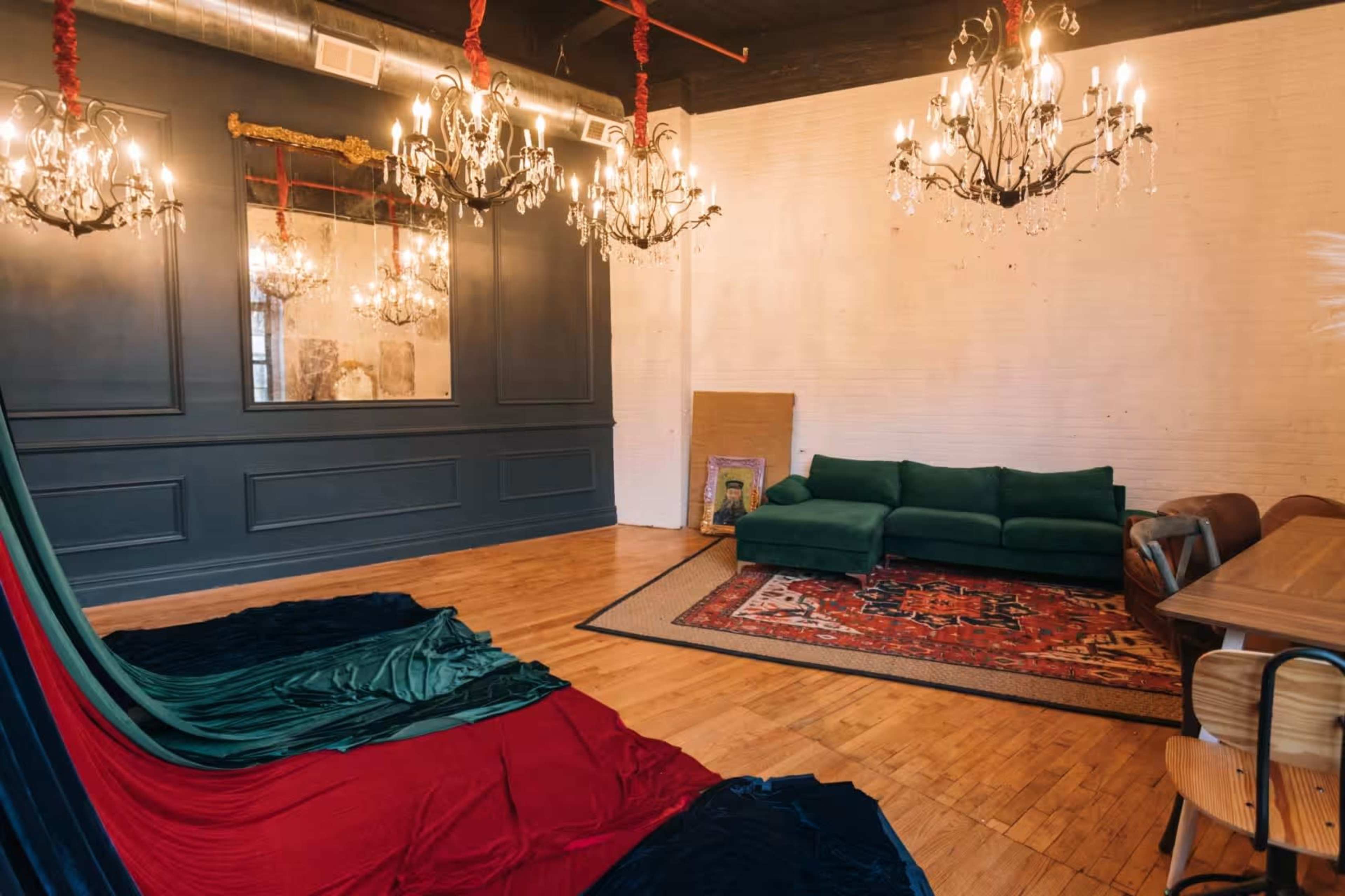 The image shows a stylish room with dark walls, several ornate chandeliers, a green sofa, a large area rug, and a colorful fabric draped on the floor.