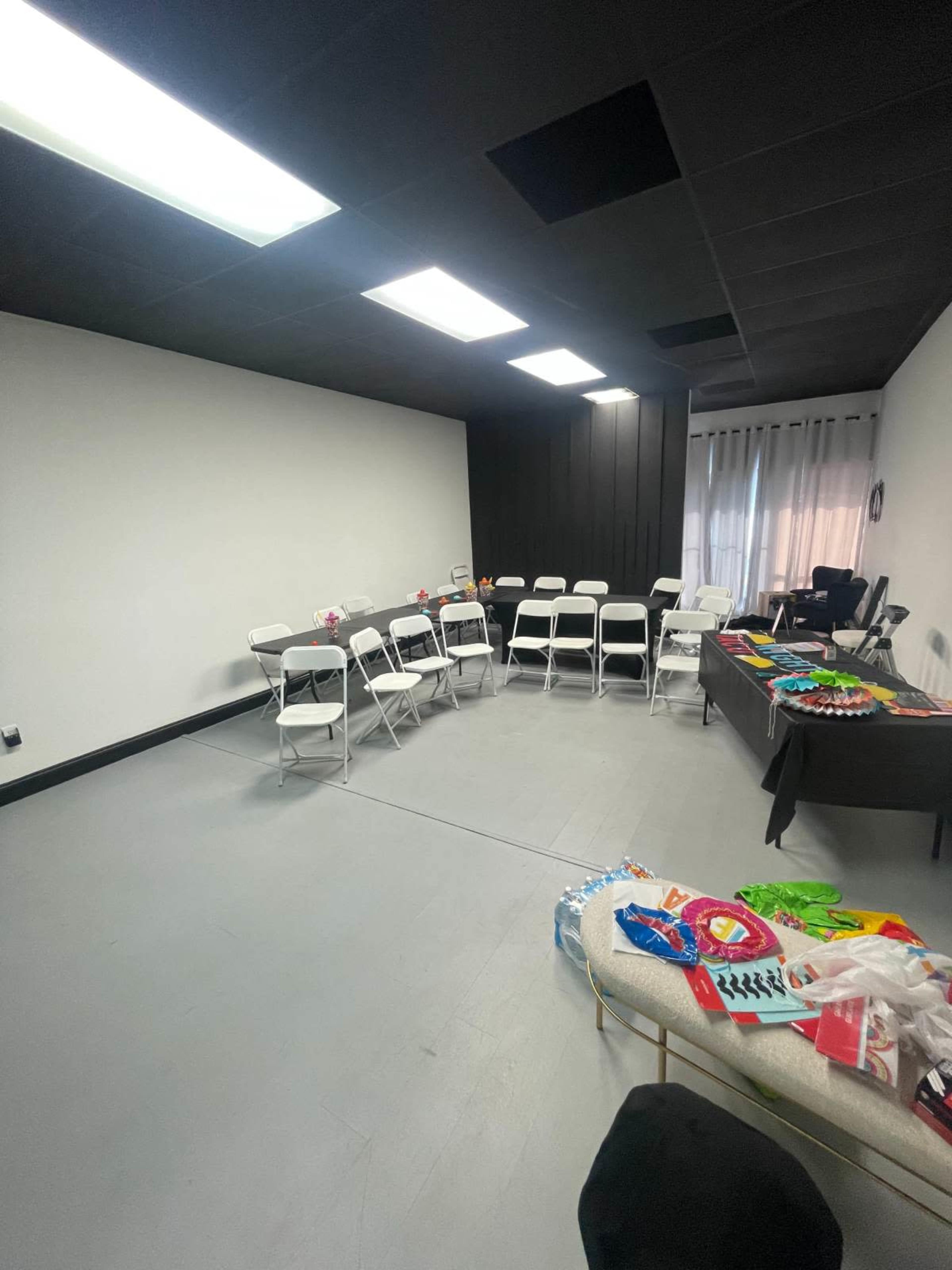 The image shows a sparsely furnished room with white walls, a few folding chairs arranged in a circle, and a table displaying colorful items.