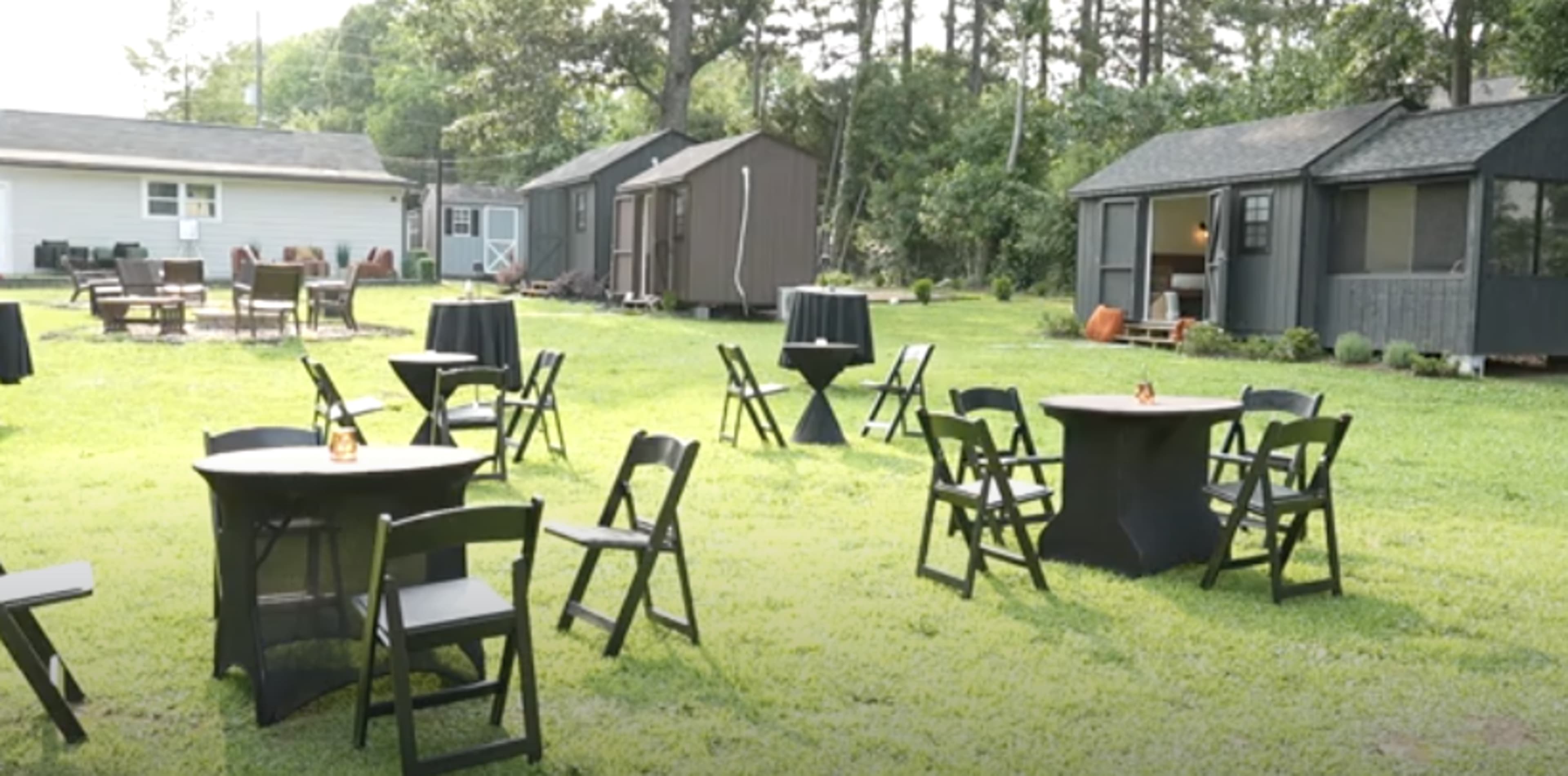 The scene shows an outdoor area with several round tables and folding chairs arranged on grassy ground, surrounded by small buildings and trees.