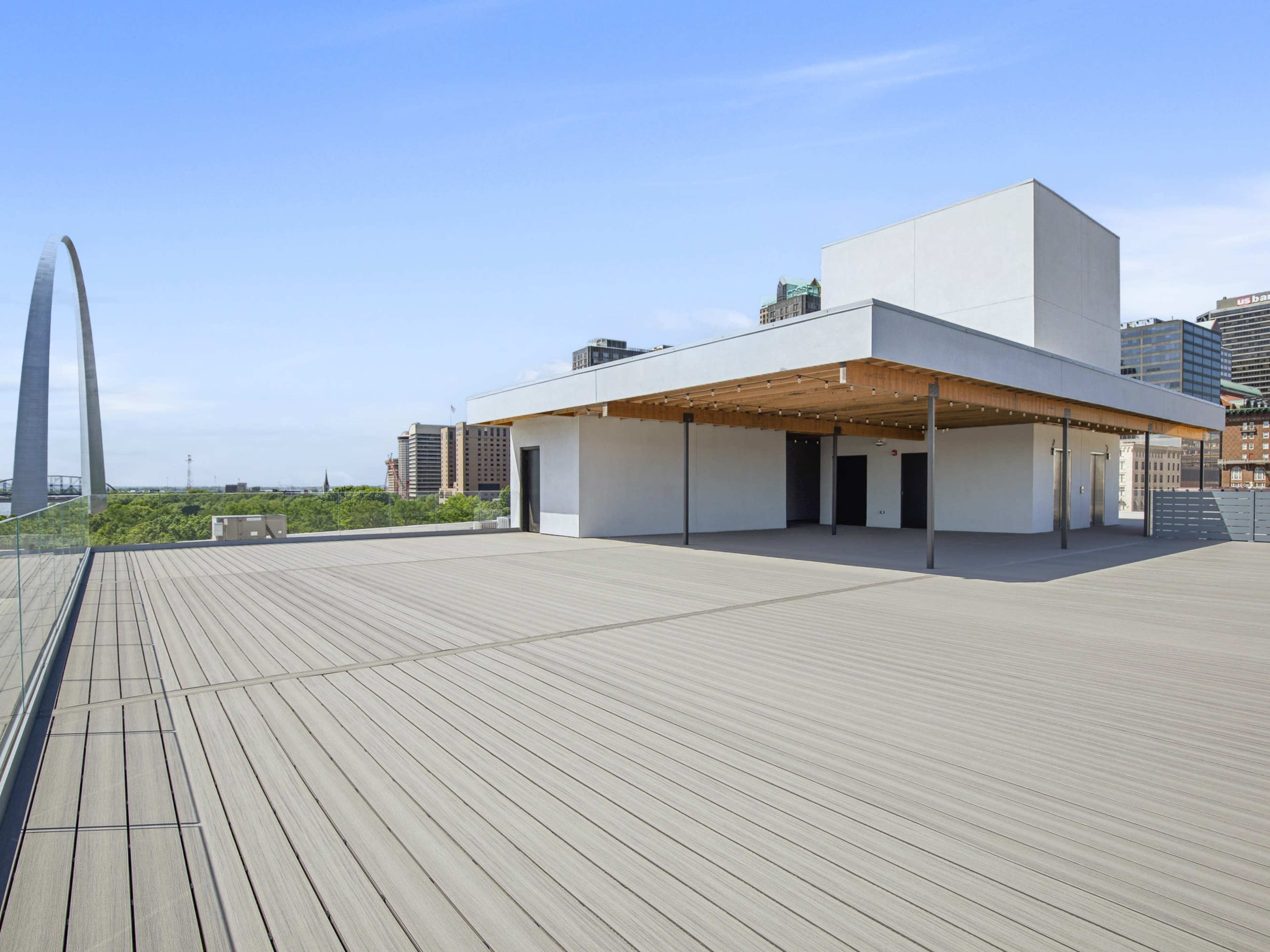 The image shows a spacious rooftop deck with a wooden floor and a modern building structure, overlooking a cityscape featuring greenery and a large arch monument in the distance.