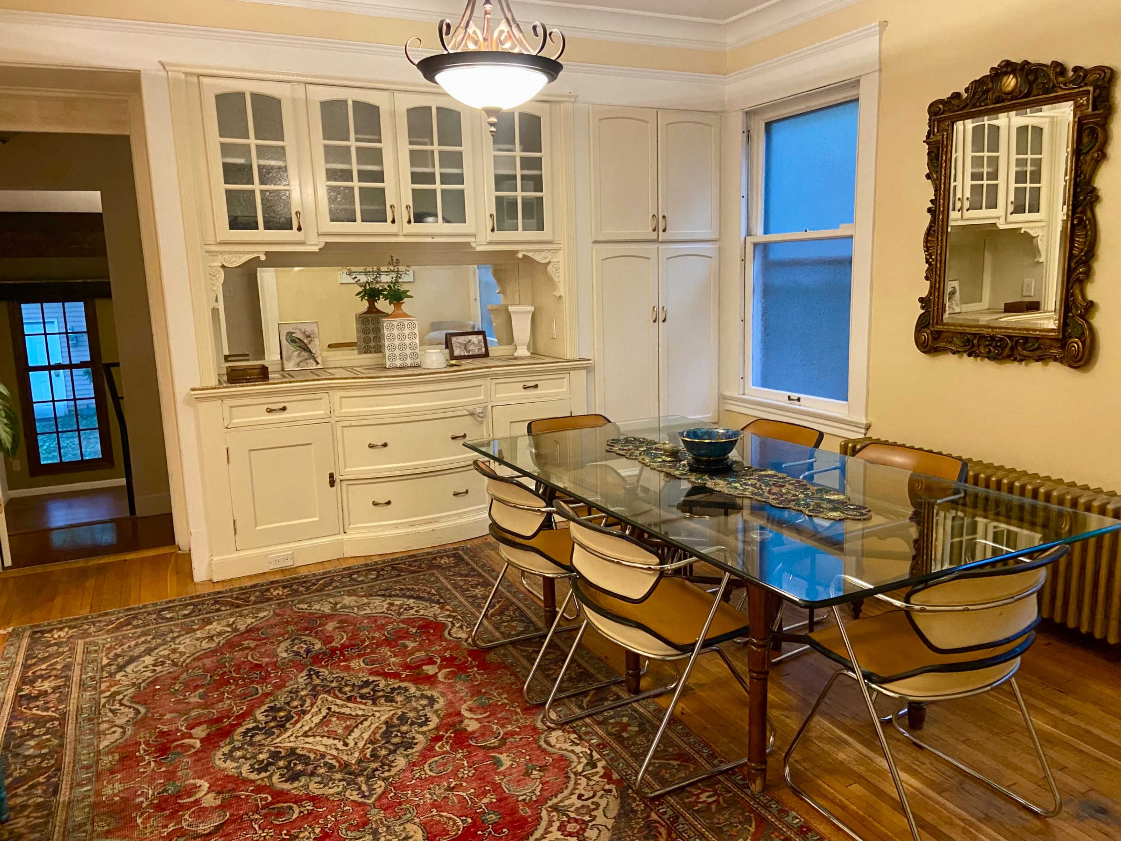 The image shows a dining room featuring a glass table surrounded by six chairs, a sideboard with decor, and a patterned rug on a hardwood floor.