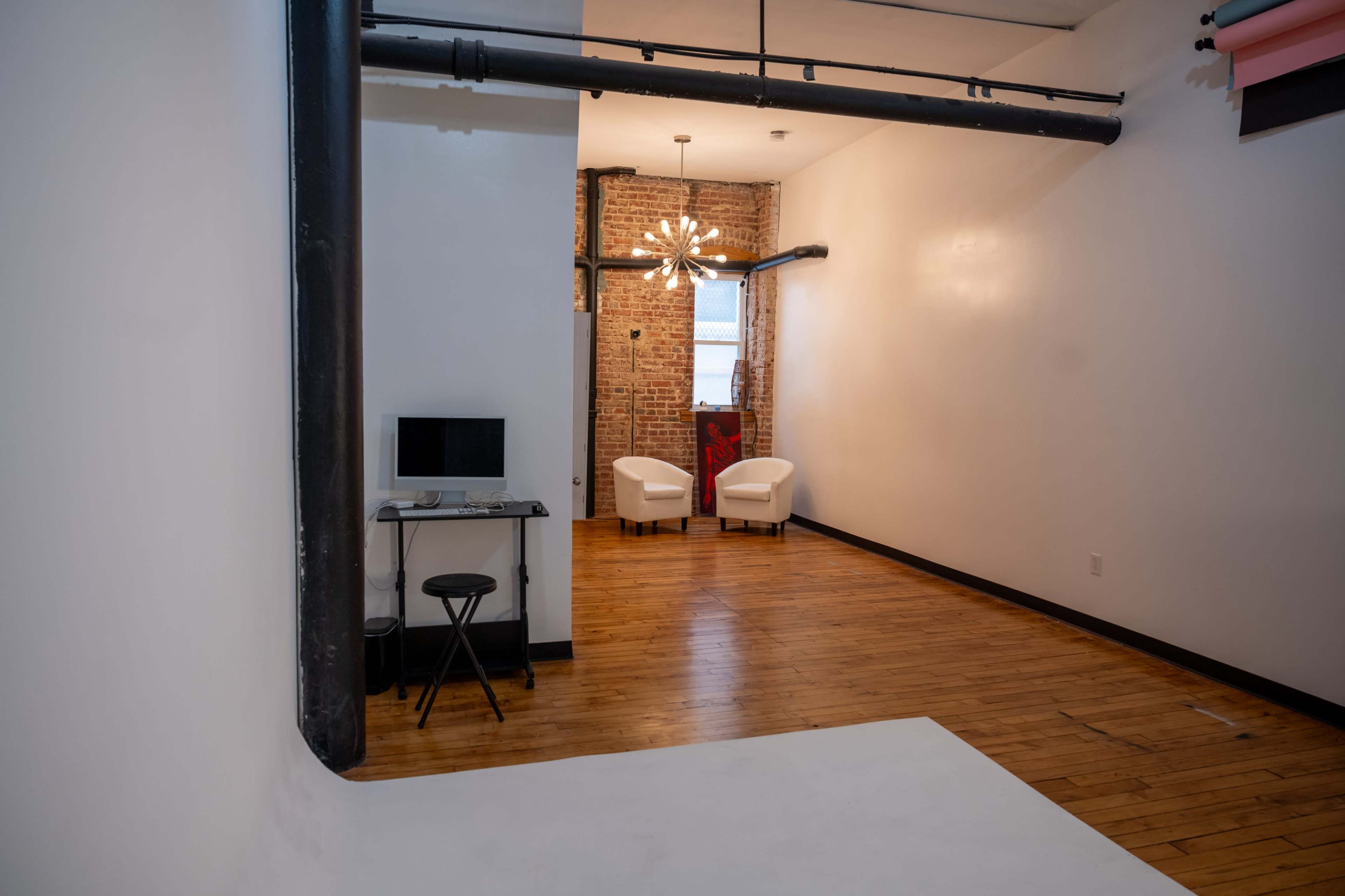 The image shows a spacious room featuring a wall with exposed brick, two white chairs facing each other, a small desk with a monitor, and wooden flooring.