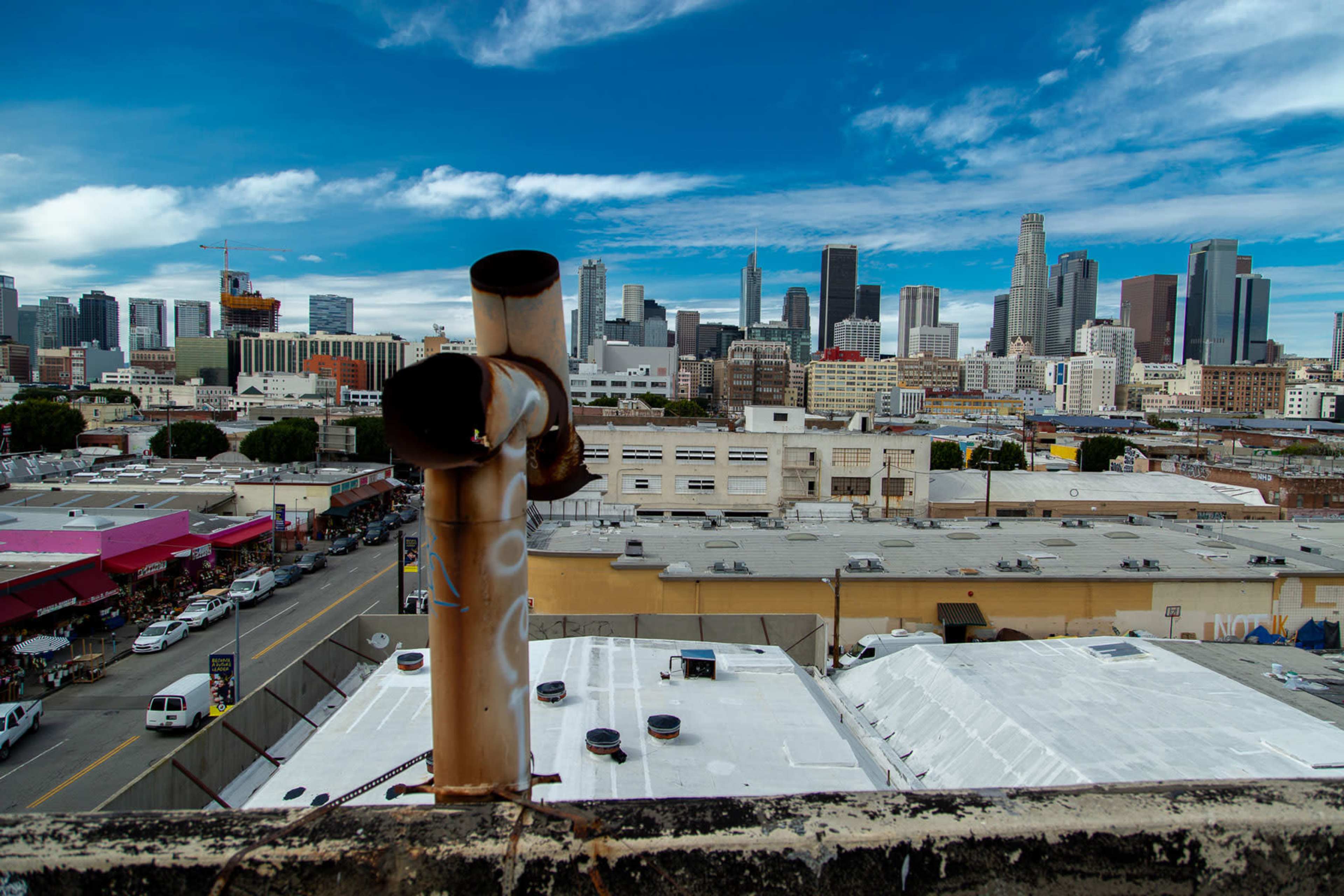 The image shows a view of the Los Angeles skyline from a rooftop, featuring buildings and a cityscape against a cloudy sky.