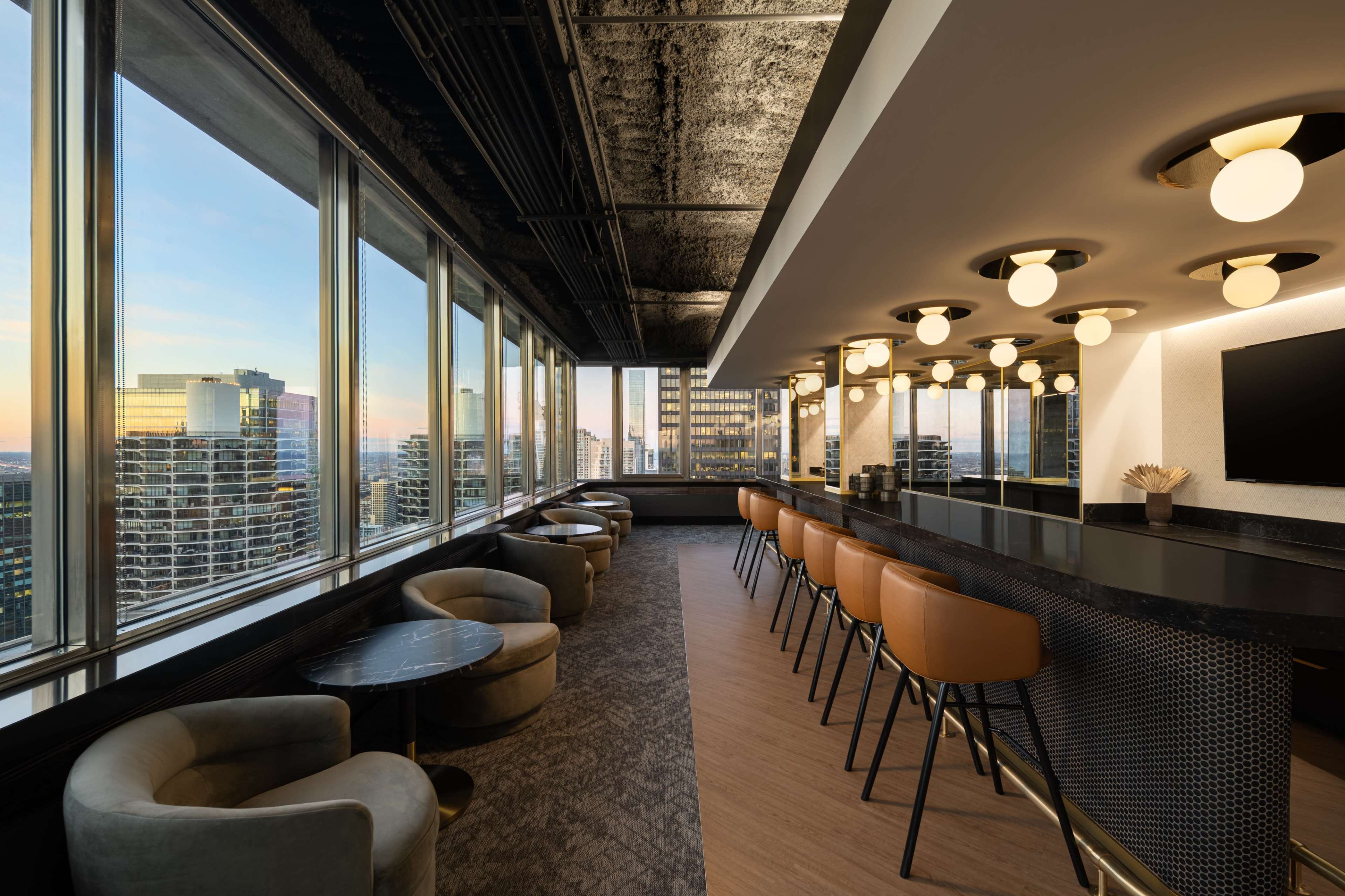 The image shows a modern bar area with high-backed stools and circular tables, featuring large windows that overlook a city skyline at sunset.