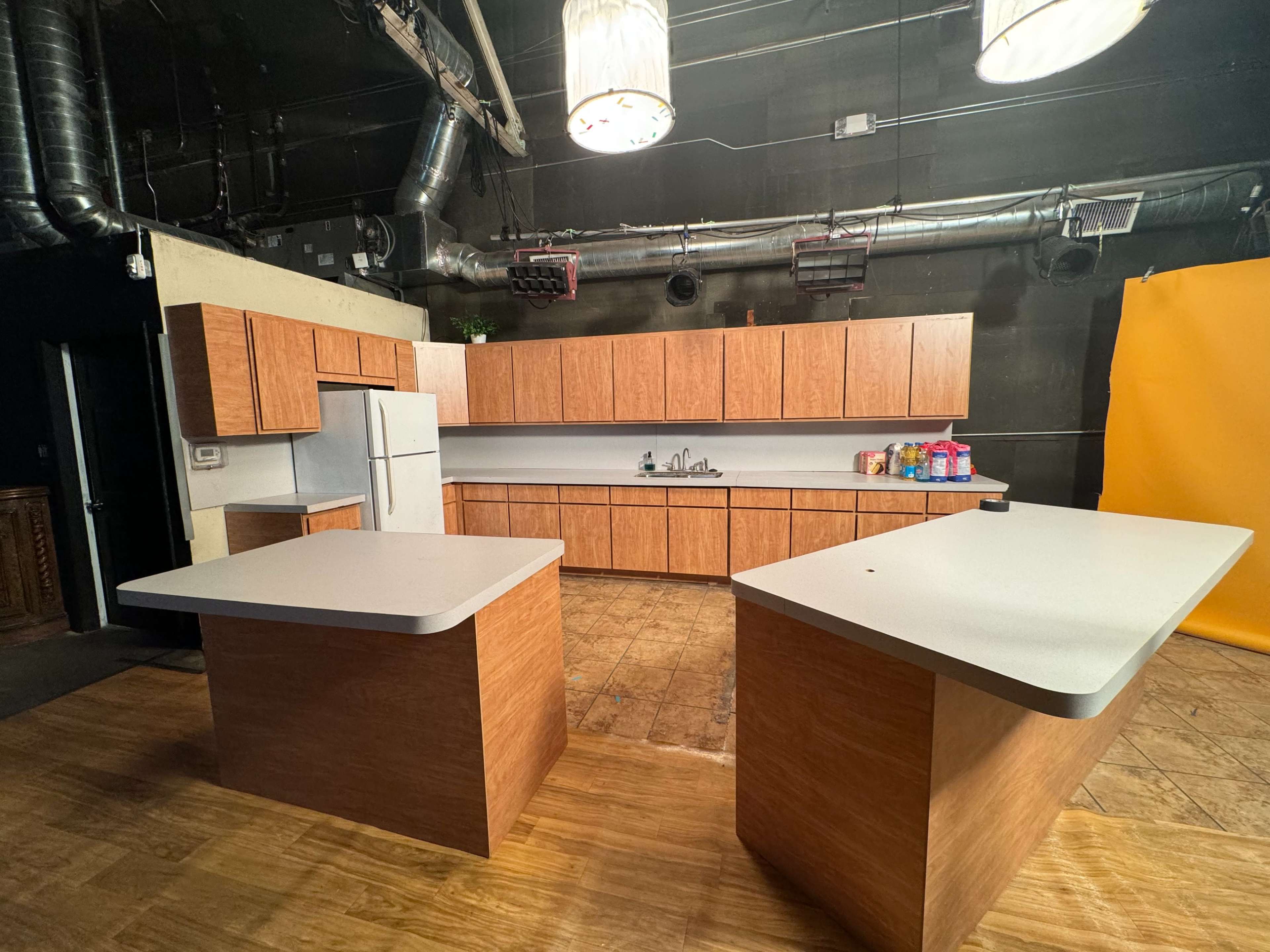 The image shows a spacious kitchen with wooden cabinetry, two large islands, and a wall-mounted refrigerator against a dark backdrop.
