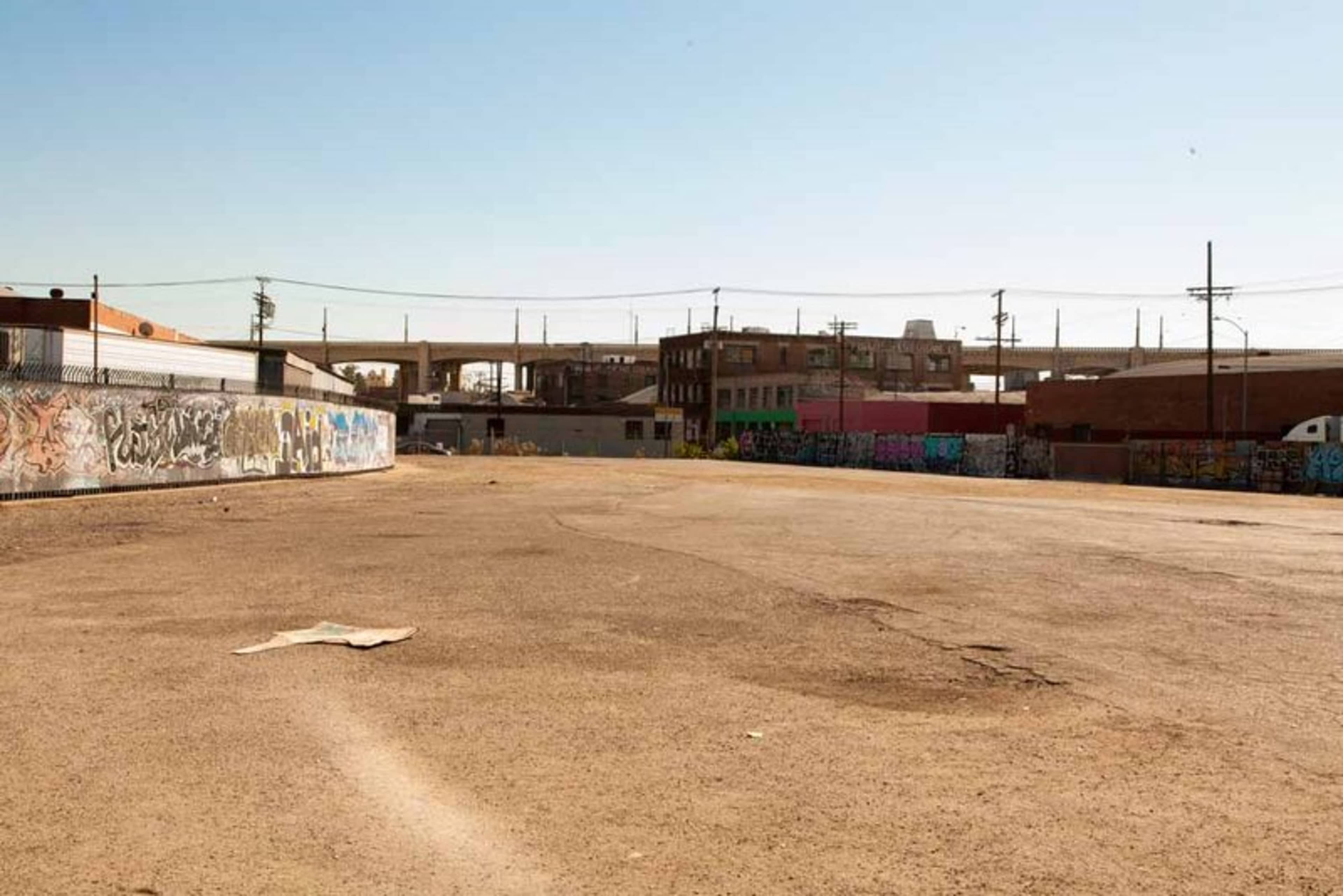The image shows an empty, gravel lot surrounded by buildings covered in graffiti, with an overpass visible in the background.