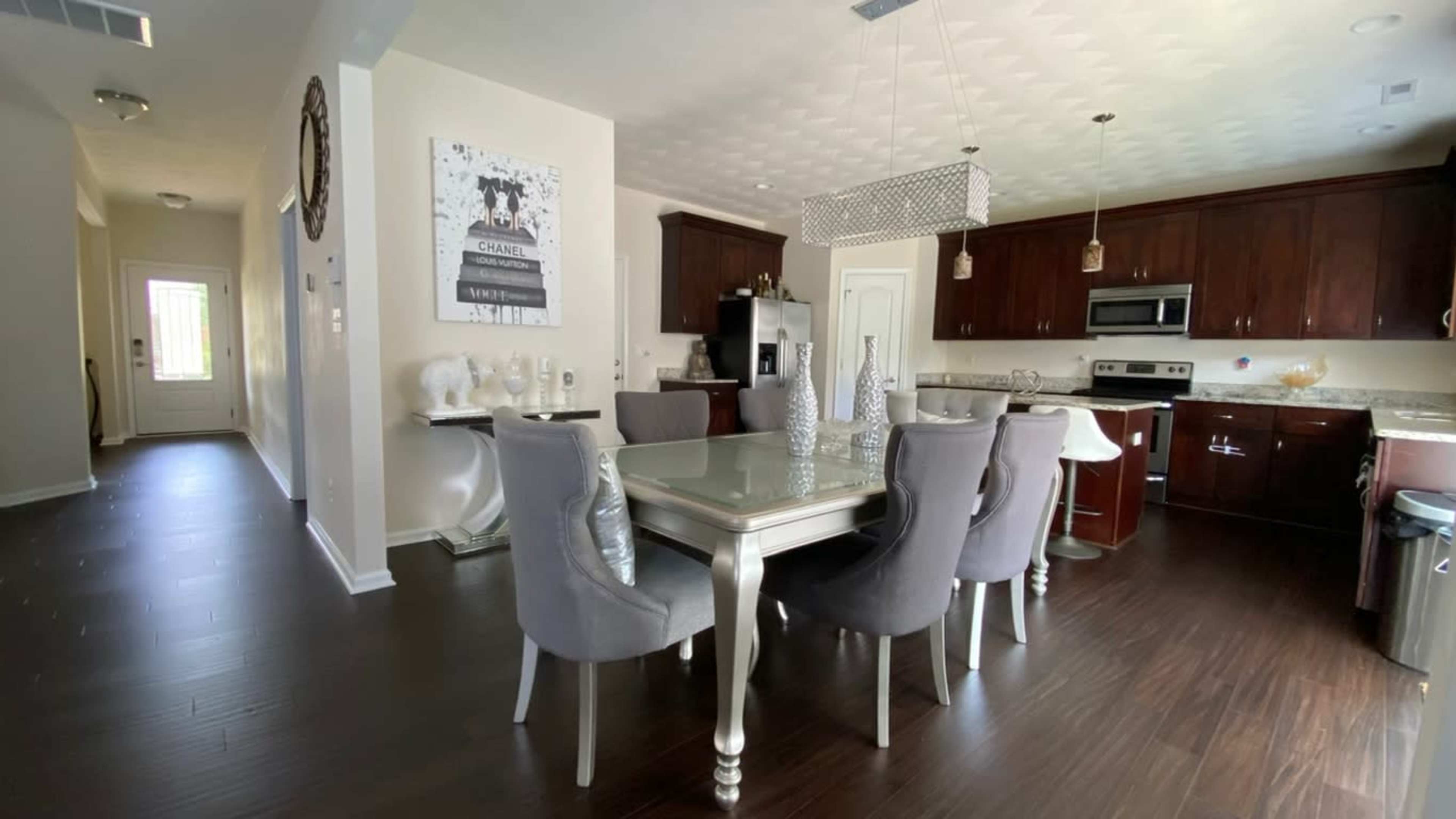 A modern kitchen and dining area with a glass-topped table surrounded by gray chairs, and wooden cabinets in the background.
